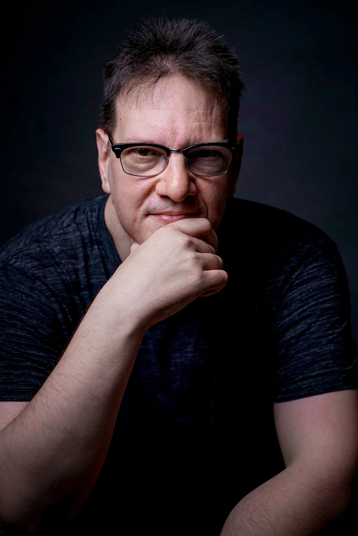 Marcus Frank Portrait des Betreibers der Webseite in einer Studioaufnahme vor dunklem Hintergrund. Die Person ist frontal zur Kamera ausgerichtet und stellt durch direkten Blickkontakt eine persönliche Verbindung zur betrachtenden Person her. Marcus sitzt leicht nach vorne geneigt und stützt das Kinn auf die Hand, wodurch eine ruhige, reflektierte und zugängliche Haltung entsteht. Diese Pose vermittelt Offenheit und lädt dazu ein, mehr über ihn zu erfahren. Er trägt ein schlichtes, dunkles T-Shirt und eine Brille mit schmalem Rahmen. Die reduzierte Kleidung und der ruhige Bildaufbau lenken den Fokus bewusst auf Gesichtsausdruck und Persönlichkeit. Das Licht ist gezielt und weich gesetzt, modelliert die Gesichtszüge klar und schafft eine ausgewogene Balance zwischen Professionalität und Natürlichkeit. Der dunkle Hintergrund sorgt für eine klare visuelle Trennung und unterstreicht die Konzentration auf die Person selbst. Der Ausdruck wirkt ruhig, aufmerksam und vertrauenswürdig. In Kombination mit der direkten Blickführung entsteht ein authentischer Eindruck, der Nähe schafft und die Funktion als persönliche Vorstellung unterstützt. Die Bildwirkung ist bewusst reduziert und charakterorientiert, um den Menschen hinter der Webseite sichtbar zu machen und Vertrauen aufzubauen. Das Portrait dient dazu, den Betreiber vorzustellen und einen ersten persönlichen Eindruck zu vermitteln.