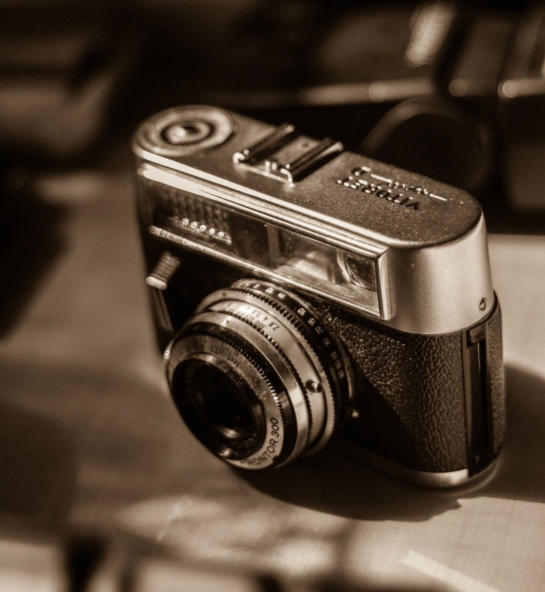 Kontakt Formular Detailaufnahme einer analogen Kompaktkamera im Vintage-Stil, fotografiert aus leicht erhöhter Perspektive mit geringer Schärfentiefe. Die Kamera ist schräg im Bild positioniert und nimmt den zentralen Fokus ein, während Vorder- und Hintergrund weich verschwimmen. Das Gehäuse besteht aus einer Kombination aus metallischen und strukturierten Oberflächen. Auf der Oberseite sind Bedienelemente sowie eine Gravur mit Marken- und Modellbezeichnung "Vitoret D Rapid" sichtbar. Die Front zeigt ein fest verbautes Objektiv mit klar erkennbaren Skalenringen und Beschriftungen. Das Licht ist weich und warm, wodurch feine Details wie Kratzer, Kanten und Materialübergänge betont werden. Reflexionen auf dem Metallgehäuse erzeugen dezente Highlights, während die Schatten sanft verlaufen. Die gesamte Aufnahme ist in einem sepiaähnlichen Farbstil gehalten, der den nostalgischen Charakter der Kamera unterstreicht und die Assoziation zu analoger Fotografie verstärkt. Der Hintergrund besteht aus unscharfen, dunklen Formen und Strukturen, die keine konkreten Elemente erkennen lassen, jedoch Tiefe erzeugen und den Blick auf das Objekt lenken. Die Bildwirkung ist ruhig und detailorientiert, mit Fokus auf Materialität, Form und fotografischer Geschichte.