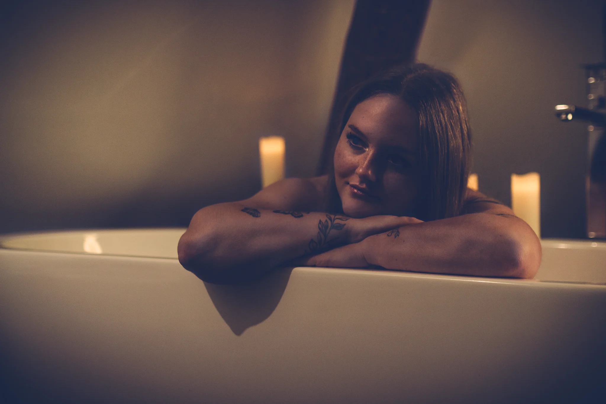 @_rebel.ig Home Shooting. Eine junge Frau liegt in einer Badewanne und stützt ihren Kopf auf die verschränkten Arme am Wannenrand. Ihr Blick ist ruhig zur Seite gerichtet, begleitet von einem sanften, entspannten Ausdruck. Mehrere Kerzen im Hintergrund spenden warmes, gedämpftes Licht, das die Szene in weiche, goldene Töne taucht und sanfte Schatten erzeugt. Dezente Details wie Armaturen und Badezimmerstruktur bleiben im Hintergrund zurückhaltend und unscharf. Die Gesamtwirkung ist ruhig, intim und von einer entspannten Wellness-Stimmung geprägt.