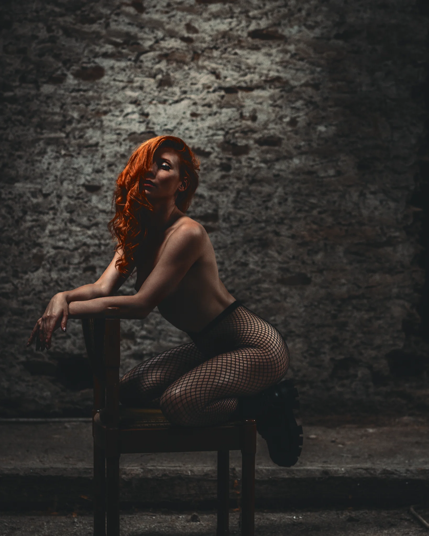 Lost Places - Low-Key-Aktporträt einer Frau mit roten Haaren in rauer, steinerner Umgebung; seitlich auf einem Stuhl sitzend und nach vorn gelehnt, wird ihr Körper durch gerichtetes Licht betont, während der dunkle, strukturierte Hintergrund eine ruhige, sinnliche und leicht dramatische Atmosphäre schafft.