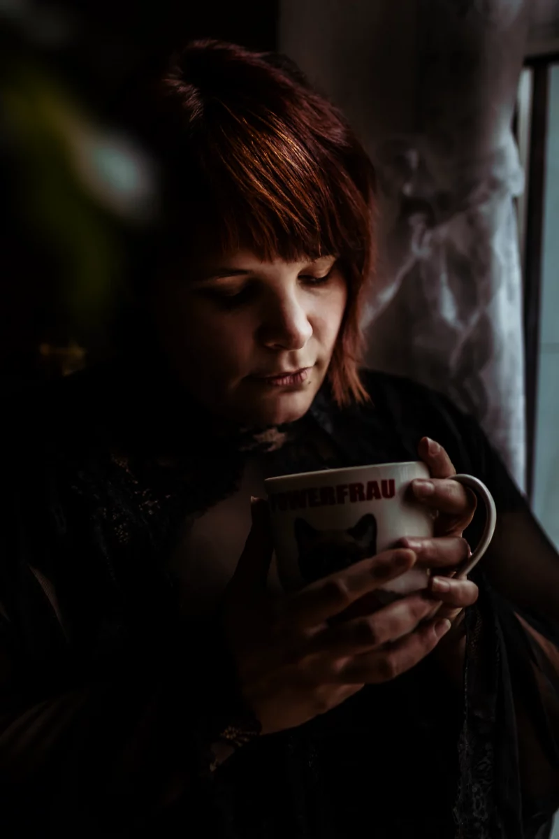 Home Shooting. Eine junge Frau mit kurzen roten Haaren sitzt nahe einem Fenster und hält eine Tasse mit beiden Händen dicht vor sich. Ihr Blick ist nach unten gerichtet, wodurch eine ruhige, leicht nachdenkliche Atmosphäre entsteht. Das Licht fällt seitlich und warm durch das Fenster ein, beleuchtet Teile ihres Gesichts und ihrer Haare und lässt den restlichen Raum in weiche Dunkelheit übergehen. Sie trägt dunkle Kleidung mit feinen, transparenten Stoffelementen, die im Licht dezent sichtbar werden. Der Hintergrund bleibt größtenteils im Schatten, wodurch der Fokus klar auf dem stillen Moment und der konzentrierten, ruhigen Haltung liegt.