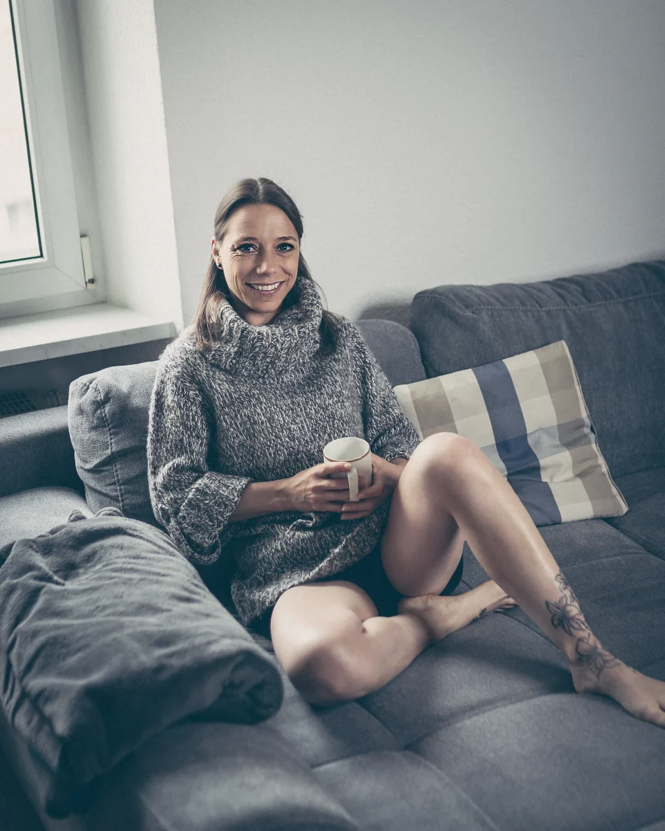 Home Shooting.Eine junge Frau sitzt locker auf einem Sofa, ein Bein angewinkelt, und hält eine Tasse mit beiden Händen. Sie blickt direkt in die Kamera und lächelt offen, wodurch eine warme, zugängliche Stimmung entsteht. Ihr grob gestrickter Pullover verstärkt den Eindruck von Gemütlichkeit, während das seitlich einfallende Tageslicht vom Fenster weiche Schatten erzeugt und die Szene natürlich ausleuchtet. Kissen und dezente Wohnraumelemente im Hintergrund bleiben unscharf und unterstreichen die ruhige, private Atmosphäre des Moments.