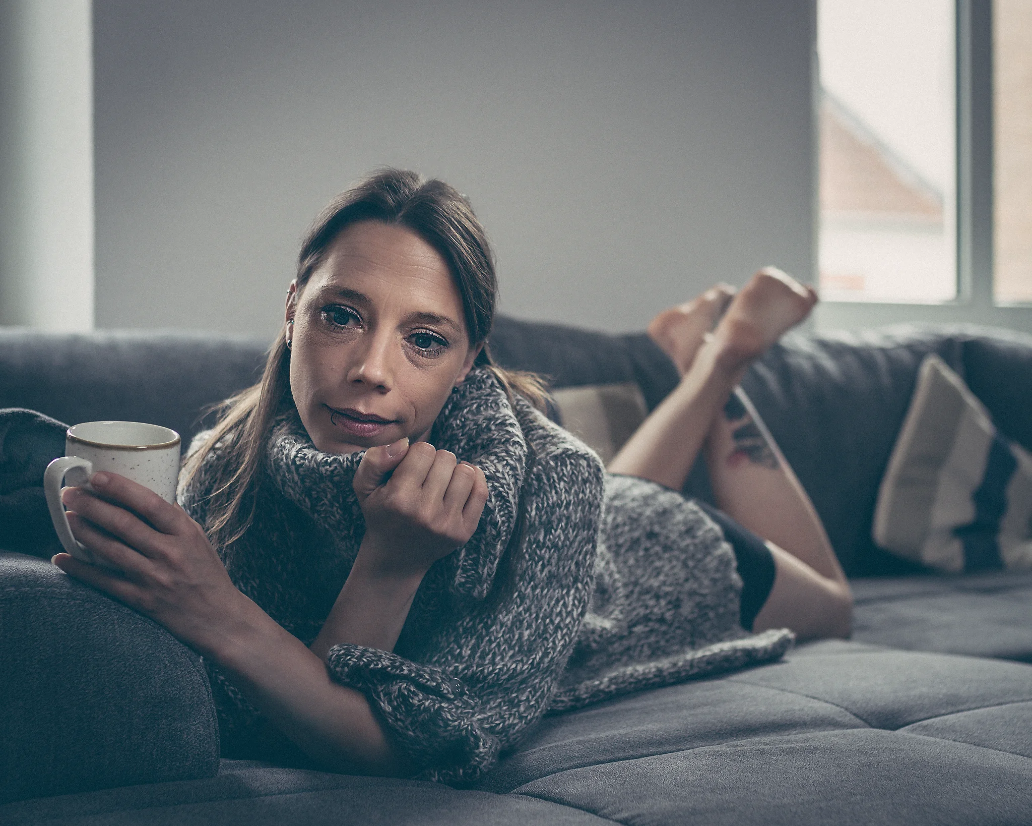 @rk_modelpage_ Home Shooting. Eine junge Frau liegt bäuchlings auf einem Sofa, den Oberkörper leicht angehoben und auf einen Arm gestützt. In der anderen Hand hält sie eine Tasse, während ihr Blick ruhig zur Seite gerichtet ist. Sie trägt einen grob gestrickten Pullover, der eine warme, gemütliche Wirkung vermittelt. Ihre Beine sind locker nach hinten ausgestreckt, wodurch eine entspannte Körperhaltung entsteht. Das Licht ist weich und kühl, fällt seitlich ein und erzeugt sanfte Schatten, die die Szene ruhig und natürlich wirken lassen. Der Hintergrund mit Fenster und Kissen bleibt unscharf und unterstreicht die private, entspannte Atmosphäre des Moments.