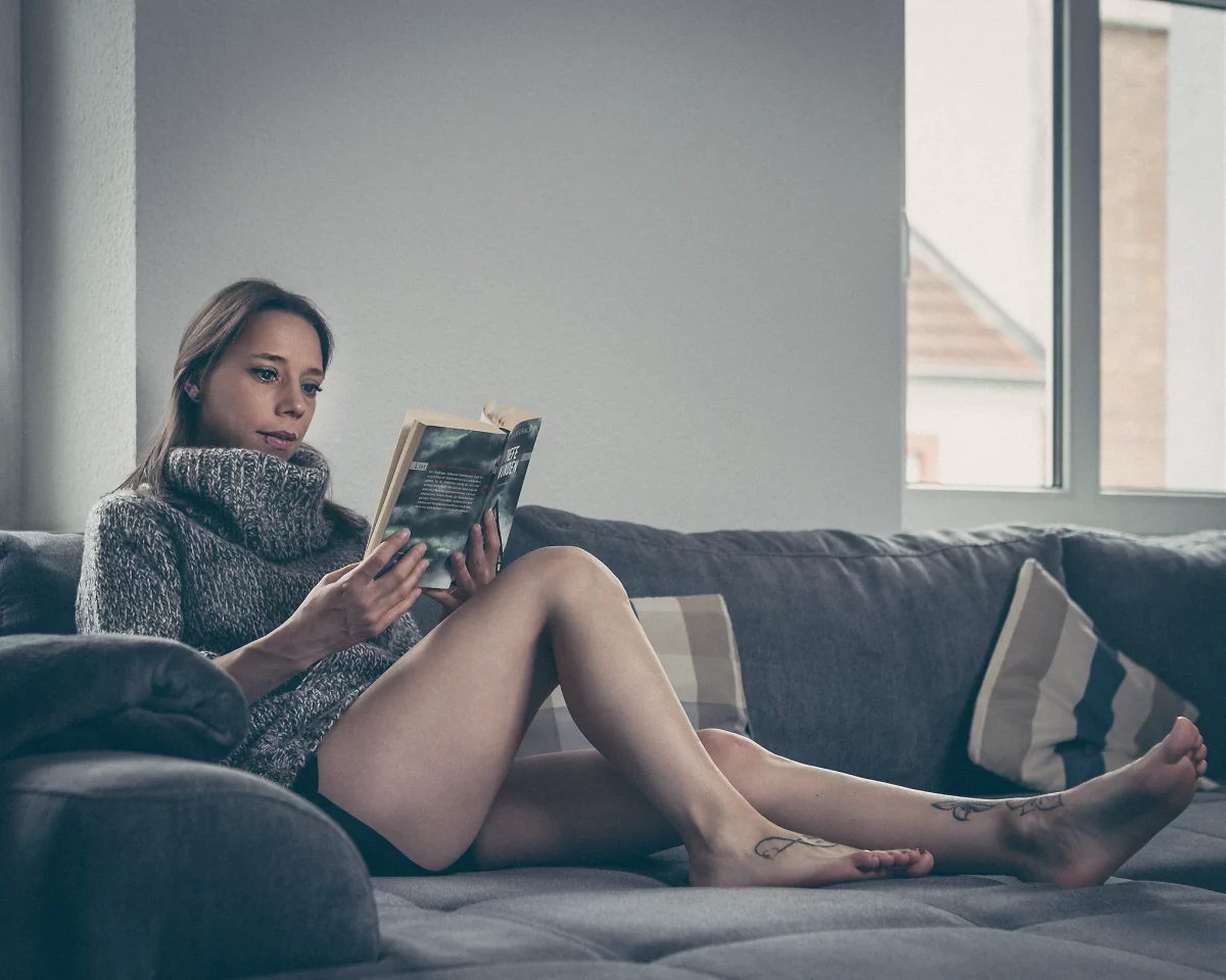Home Shooting.Eine junge Frau sitzt bequem auf einem Sofa, die Beine ausgestreckt und leicht angewinkelt, während sie ein aufgeschlagenes Buch in den Händen hält. Ihr Blick ist konzentriert auf die Seiten gerichtet, wodurch eine ruhige, in sich gekehrte Stimmung entsteht. Sie trägt einen lockeren, grob gestrickten Pullover, der den Eindruck von Wärme und Gemütlichkeit verstärkt. Das Licht ist weich und kühl, fällt von der Seite ein und erzeugt sanfte Schatten. Im Hintergrund sind ein Fenster und schlichte Wohnraumelemente zu erkennen, die jedoch unscharf bleiben und die entspannte, private Atmosphäre des Moments unterstreichen.