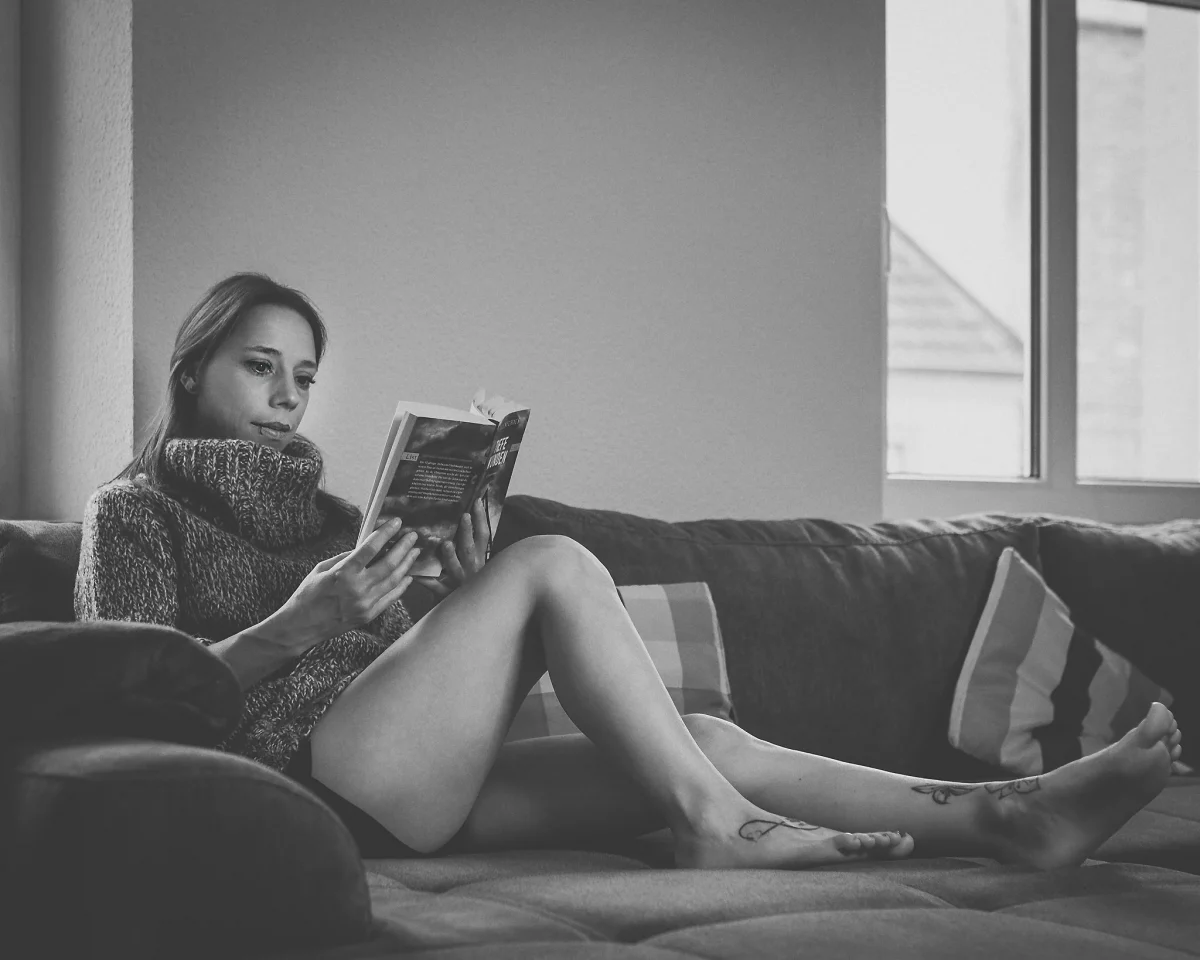 Home Shooting.Eine junge Frau sitzt bequem auf einem Sofa, die Beine ausgestreckt und leicht angewinkelt, während sie ein aufgeschlagenes Buch in den Händen hält. Ihr Blick ist konzentriert auf die Seiten gerichtet, wodurch eine ruhige, in sich gekehrte Stimmung entsteht. Sie trägt einen lockeren, grob gestrickten Pullover, der den Eindruck von Wärme und Gemütlichkeit verstärkt. Das Licht ist weich und kühl, fällt von der Seite ein und erzeugt sanfte Schatten. Im Hintergrund sind ein Fenster und schlichte Wohnraumelemente zu erkennen, die jedoch unscharf bleiben und die entspannte, private Atmosphäre des Moments unterstreichen.