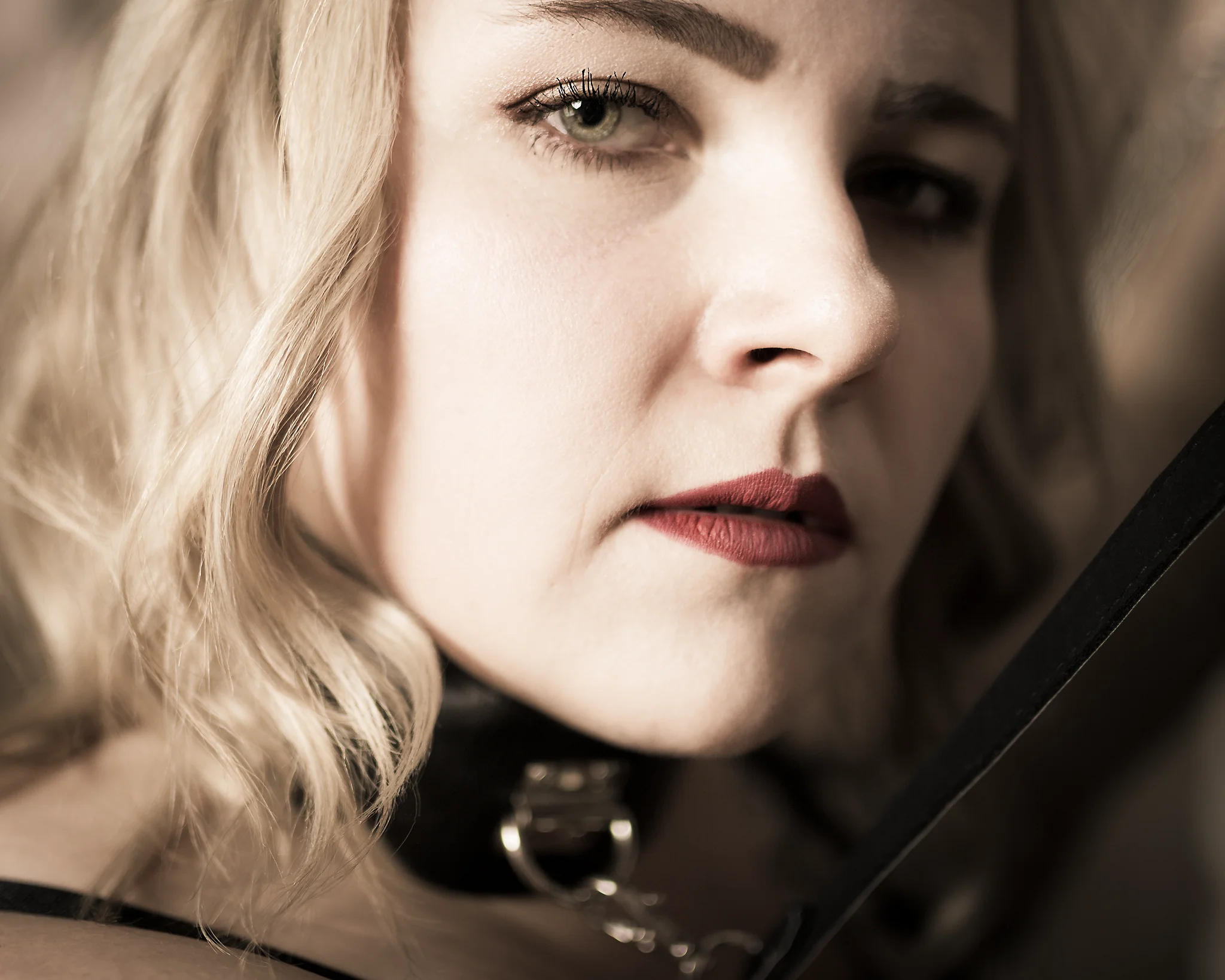 Portrait - Nahaufnahme eines Gesichts mit hellem, welligem Haar und dezentem Make-up, die Person trägt ein schwarzes Halsband mit Metallring; der Blick ist ruhig und direkt, das Licht weich und warm mit geringer Tiefenschärfe.