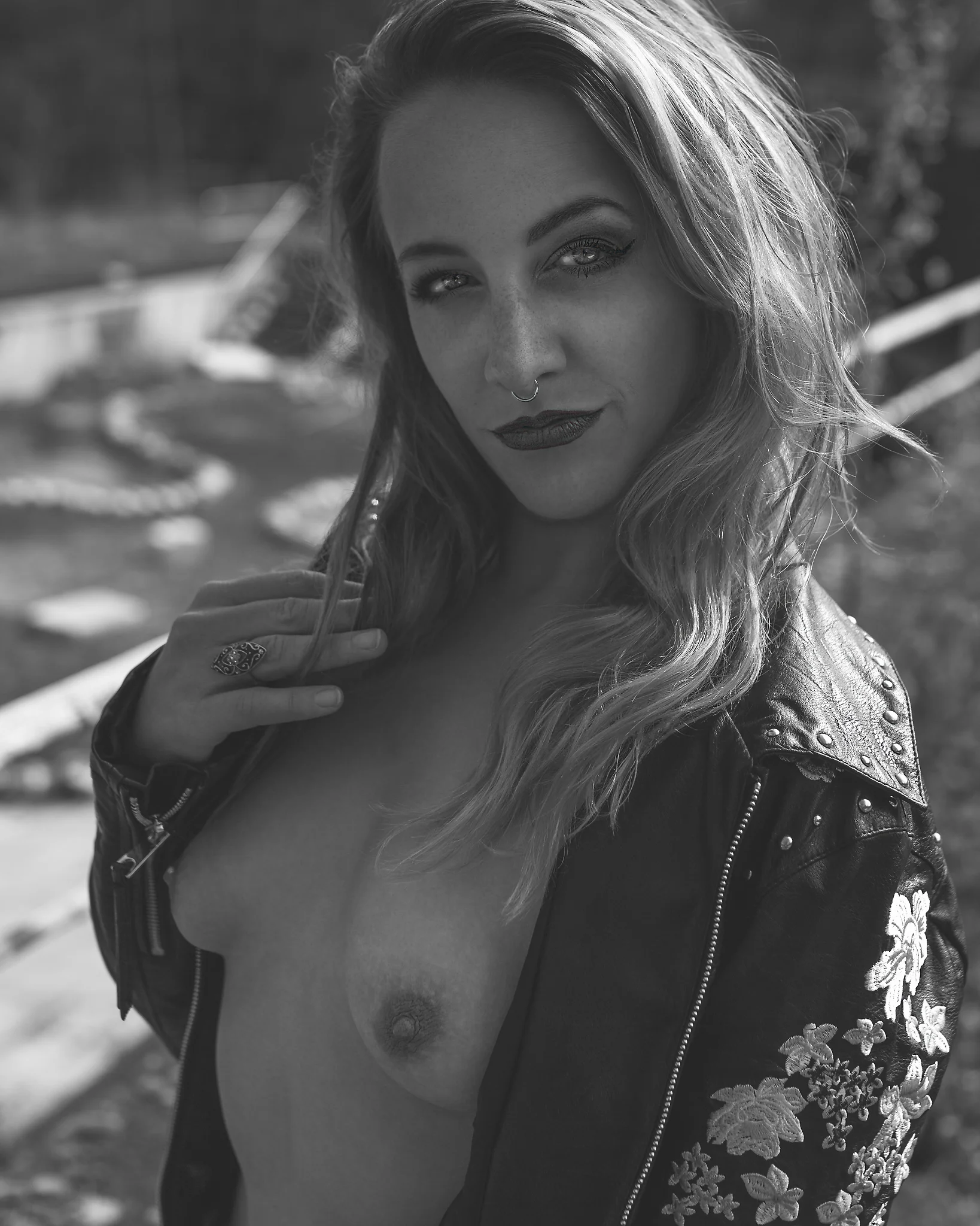 Portrait - Schwarzweiß-Porträt einer Person im Außenbereich mit offener Lederjacke und unbekleidetem Oberkörper, leicht gedrehte Haltung und direkter Blick in die Kamera; weiches natürliches Licht und geringe Tiefenschärfe betonen Gesichtszüge, Hautstruktur und Kontraste, während der Hintergrund ruhig in Unschärfe bleibt.