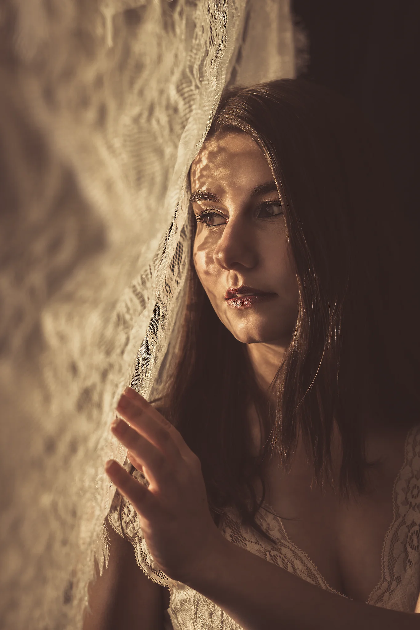 Portrait - Porträt einer Person mit langen dunklen Haaren in ruhiger, nach innen gerichteter Haltung, seitlich an einem halbtransparenten Vorhang; warmes, gerichtetes Licht fällt durch das Gewebe und zeichnet weiche Schattenmuster ins Gesicht, geringe Tiefenschärfe und sanfte Kontraste betonen Hautstruktur, Blickrichtung und die ruhige, intime Stimmung des Bildes.