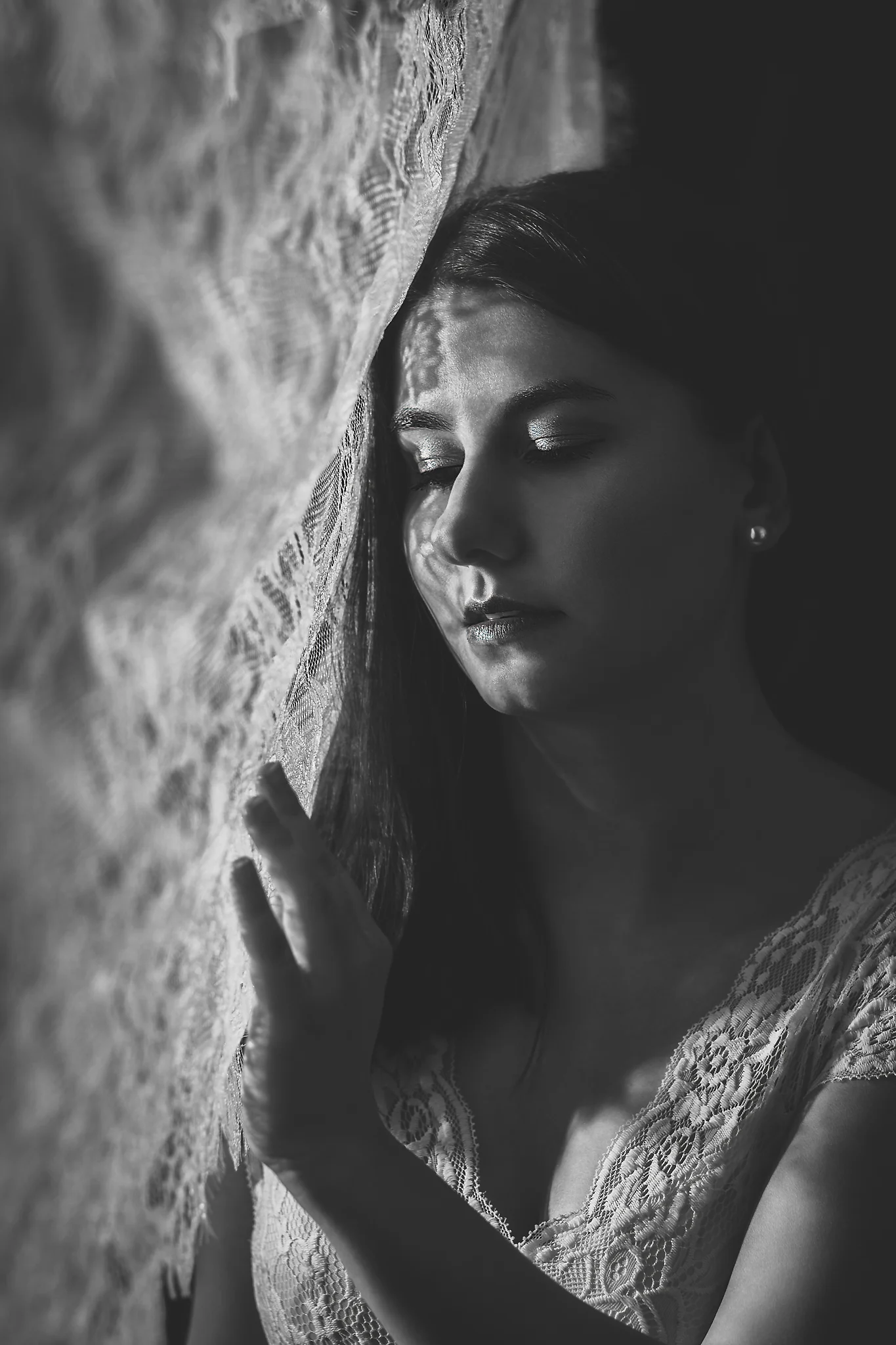 Portrait - Schwarzweiß-Porträt einer Person mit langen dunklen Haaren in ruhiger, nach innen gerichteter Haltung, seitlich an einem strukturierten Vorhang angelehnt; sanftes Licht fällt durch das Gewebe und erzeugt weiche Schattenmuster im Gesicht, geringe Tiefenschärfe und dezente Kontraste unterstreichen die stille, intime Bildwirkung.