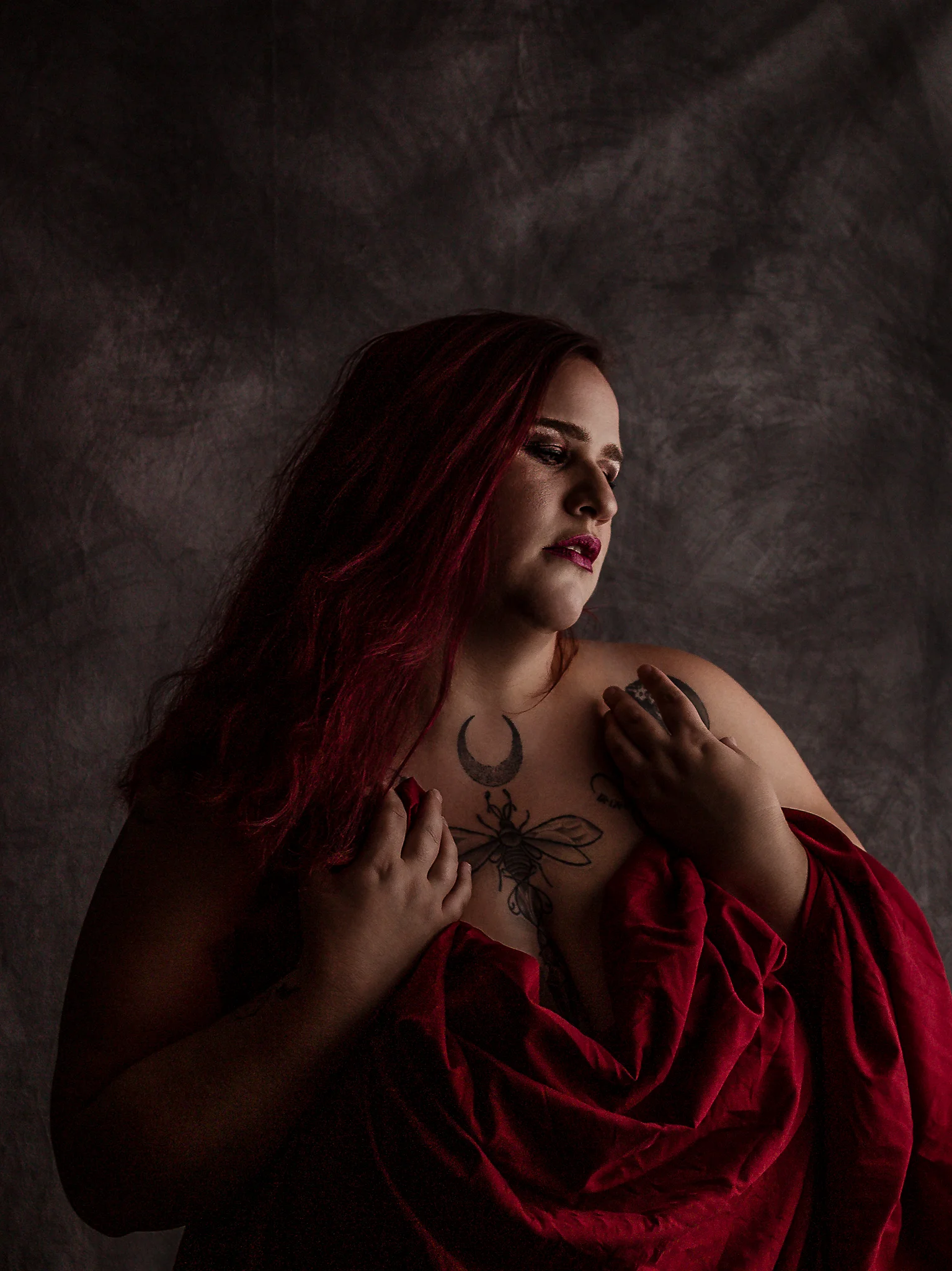 Portrait - Porträt einer Person mit langem rotem Haar, in ruhiger, leicht abgewandter Haltung, der Oberkörper nur teilweise von einem roten Tuch bedeckt; weiches, gerichtetes Licht modelliert Gesicht und Körper und hebt Hautstruktur sowie Tattoos hervor, während der dunkle, strukturierte Hintergrund in Unschärfe bleibt und eine warme, intime Bildwirkung erzeugt.