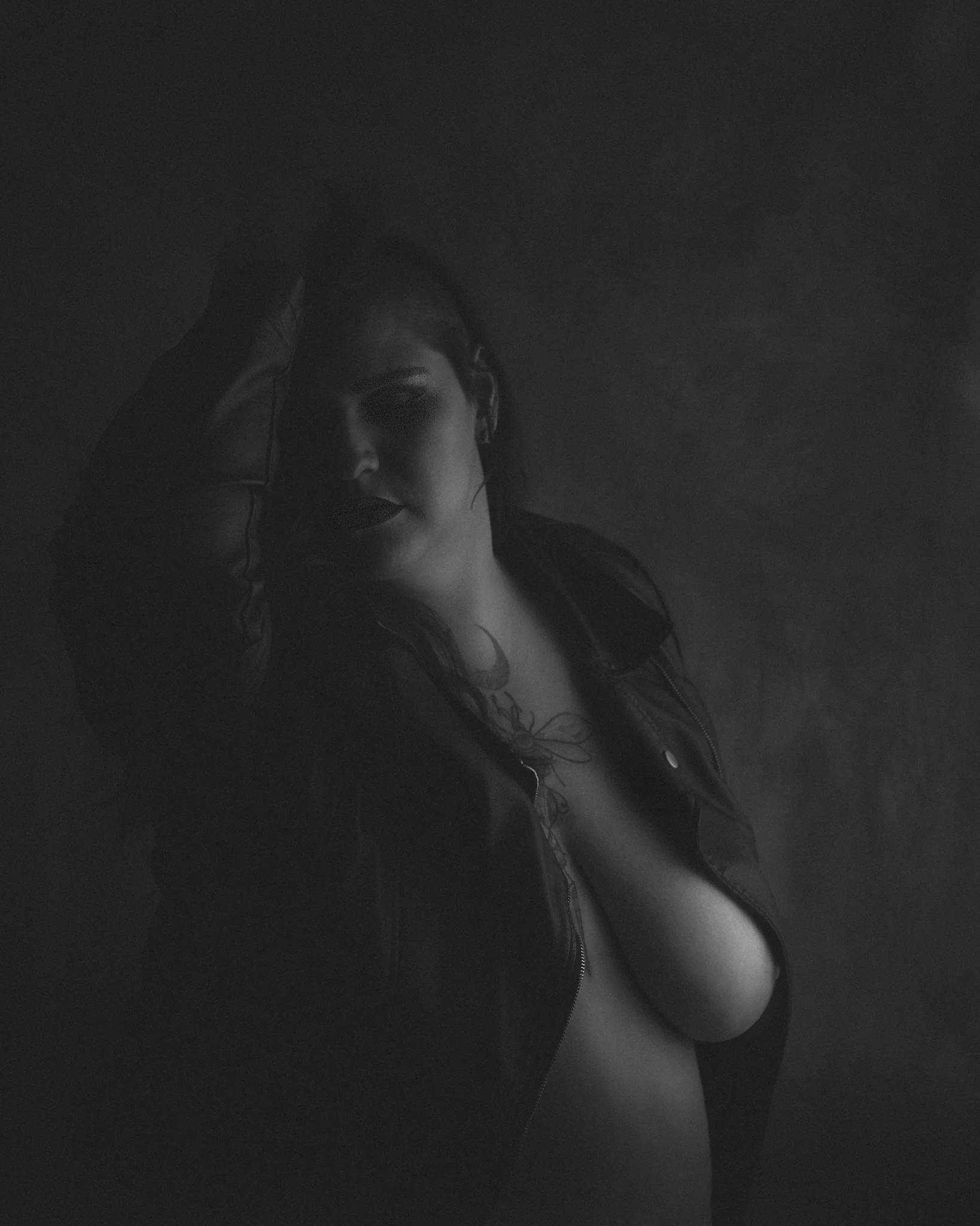 Portrait - Schwarzweiß-Porträt einer Person mit teilweise geöffneter Jacke und unbekleidetem Oberkörper in ruhiger, leicht gedrehter Haltung; der Blick seitlich aus dem Bild geführt, weiches, sehr zurückhaltendes Licht modelliert Körper und Gesicht nur partiell und lässt große Bereiche im Schatten, wodurch eine reduzierte, intime und geheimnisvolle Bildwirkung entsteht.