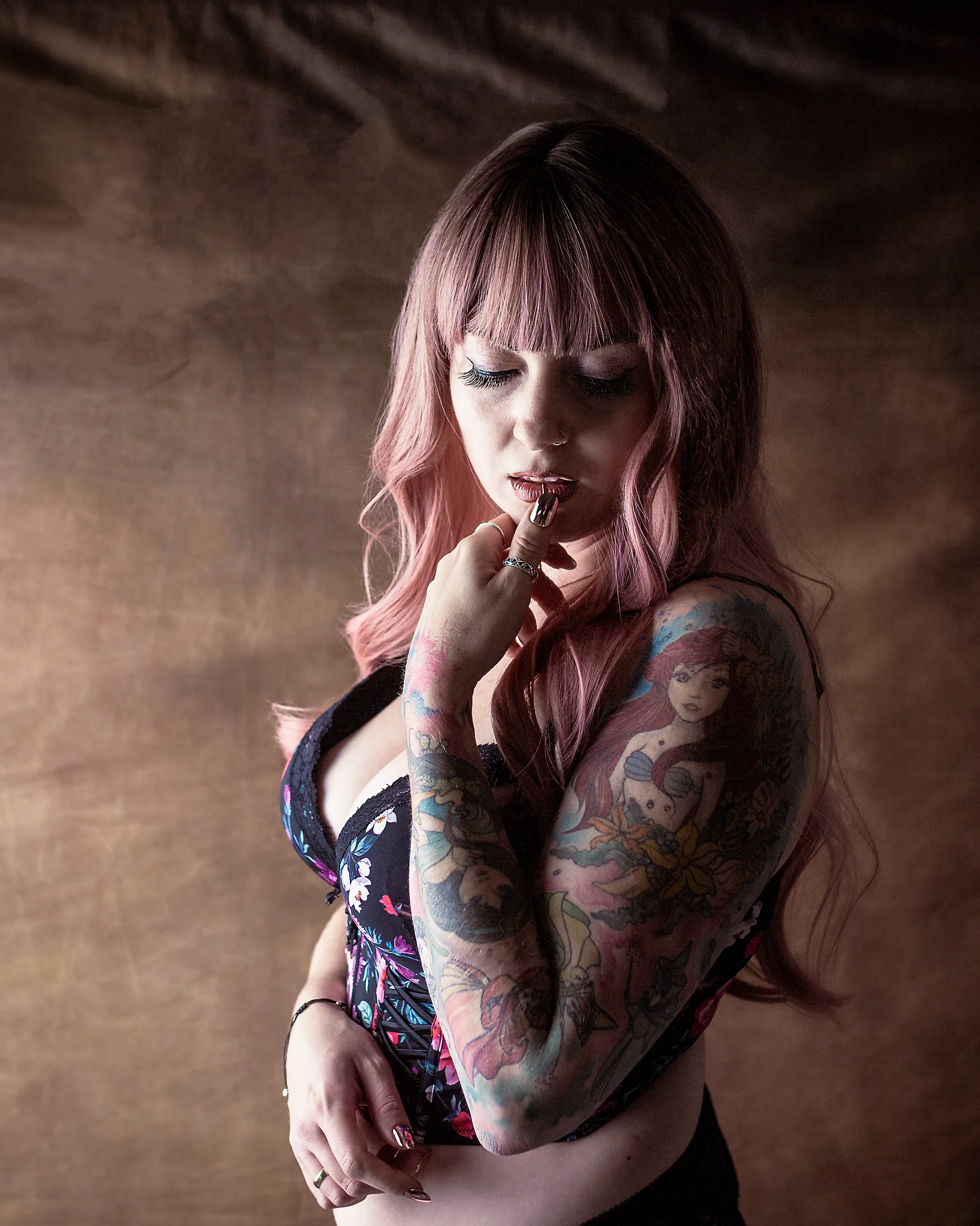 Portrait - Porträt einer Person mit langem, rosafarbenem Haar und tätowiertem Arm, in leicht gedrehter Haltung mit der Hand am Mund; ein eng anliegendes Oberteil betont die Figur, während weiches, gerichtetes Licht Gesicht und Haut modelliert und die Farben der Tattoos hervorhebt. Der warme, strukturierte Hintergrund bleibt dezent unscharf und unterstützt die ruhige, sinnliche Bildwirkung.