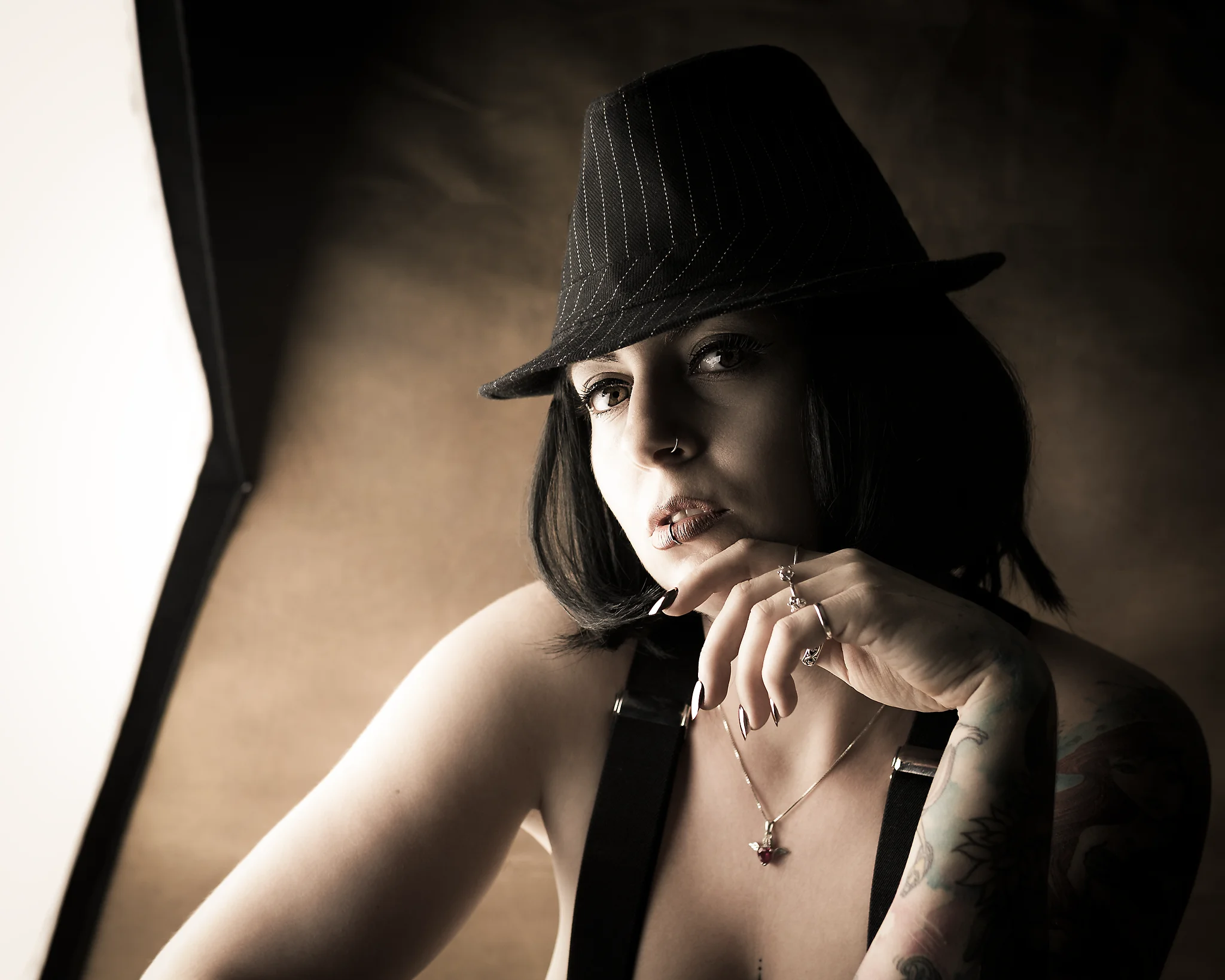 Portrait - Porträt einer Person mit dunklem, kinnlangem Haar und Hut, in nachdenklicher Haltung mit leicht erhobener Hand am Kinn; weiches Seitenlicht aus einer Softbox modelliert Gesicht und Oberkörper und betont Hauttöne sowie feine Details wie Schmuck und Tattoos, während der warme, zurückhaltende Hintergrund dezent in Unschärfe verläuft und eine ruhige, intime Studioatmosphäre entsteht.