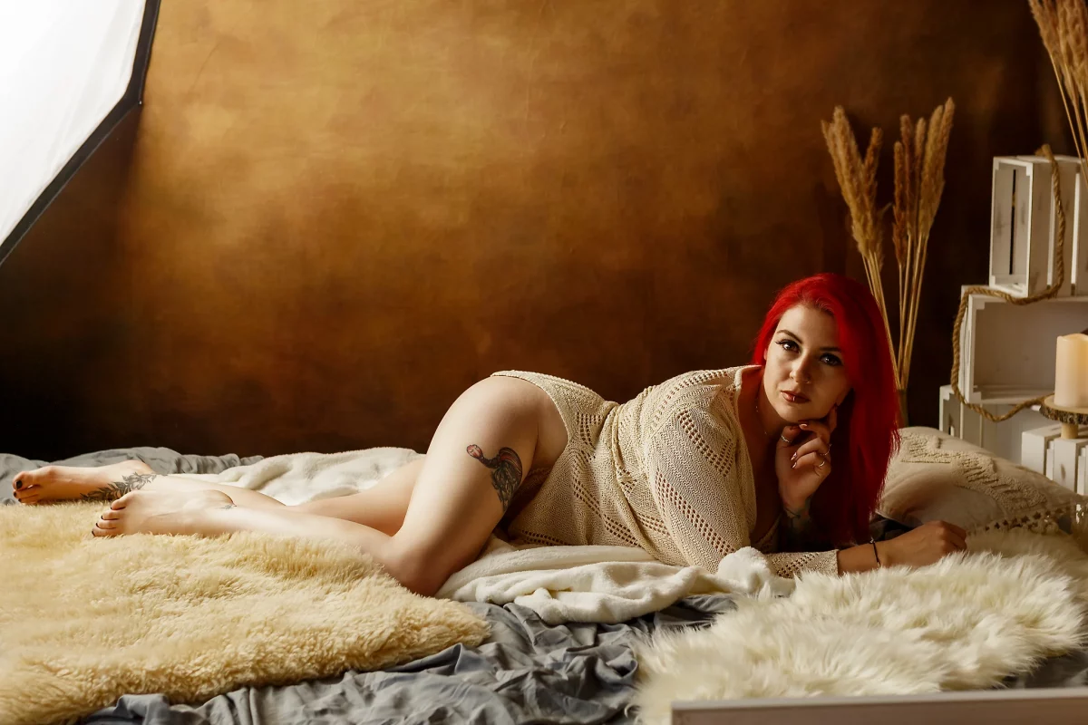 Lingerie Shooting im Studio. Dessousfotografie einer Frau in liegender Pose auf einem Bett mit weichen Decken und Fellen vor einem warm getönten Studiohintergrund. Die Frau liegt seitlich ausgestreckt, der Körper leicht in einer S-Linie geformt, ein Bein angewinkelt, das andere lang nach hinten geführt. Ein Arm stützt den Oberkörper, während die andere Hand locker am Kinn liegt. Sie trägt ein helles, grob gestricktes Oberteil, das den Körper nur teilweise bedeckt. Der Blick ist direkt zur Kamera gerichtet und erzeugt eine ruhige, präsente Ausstrahlung. Auffällige rote Haare setzen einen starken Farbkontrast zur warmen, erdigen Umgebung. Das Licht ist weich und seitlich geführt, modelliert die Körperkonturen und hebt die unterschiedlichen Materialstrukturen von Stoff, Fell und Haut hervor. Dekorative Elemente wie Kissen, Holzkisten und Trockenpflanzen ergänzen die Szene, bleiben jedoch dezent im Hintergrund. Die Kombination aus liegender Pose, direktem Blick und harmonischer Farbgestaltung erzeugt eine sinnliche, ruhige Bildwirkung innerhalb der Fine-Art-Dessousfotografie.