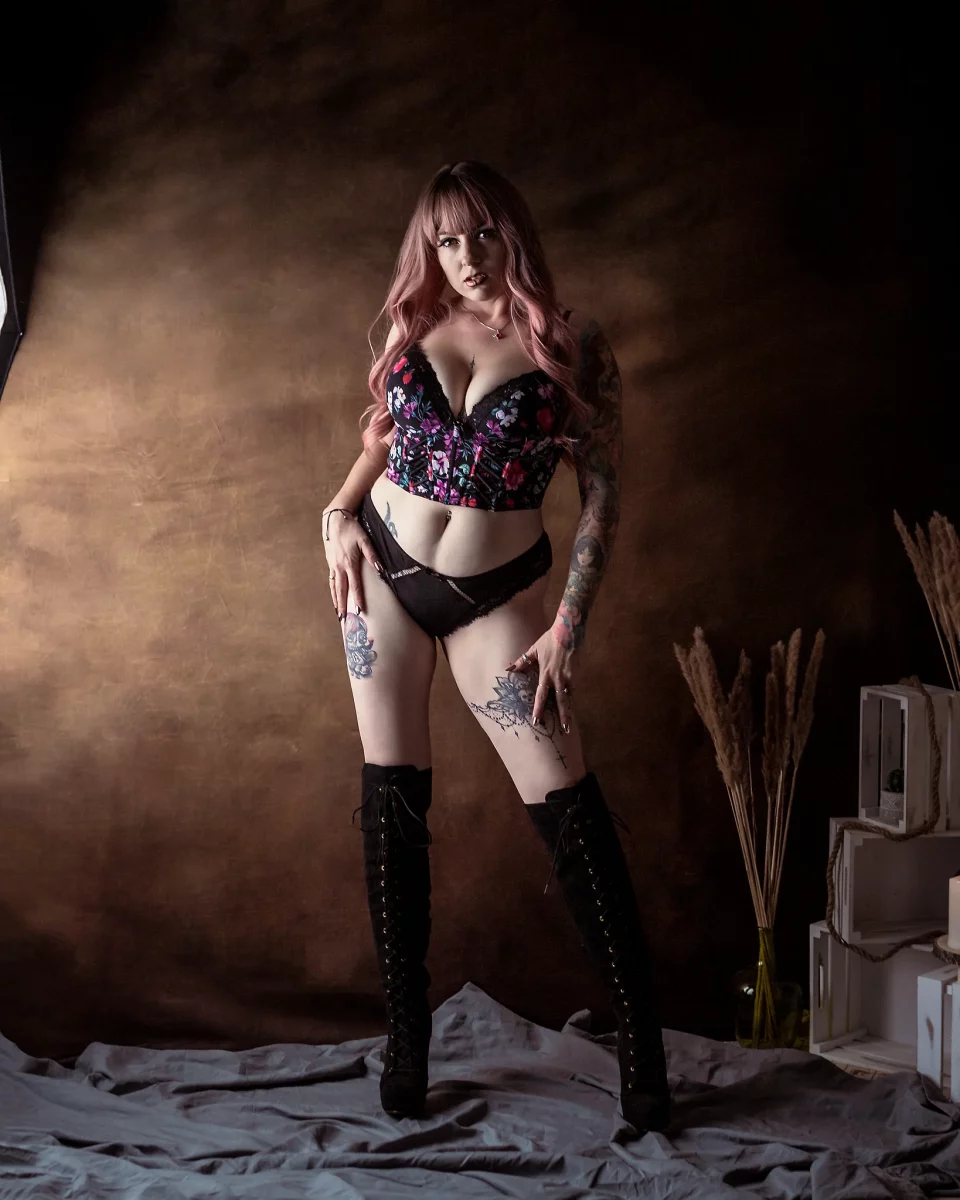 Lingerie Shooting im Studio. Dessousfotografie einer Frau in stehender Pose vor einem warm getönten, strukturierten Studiohintergrund. Die Frau steht frontal mit leicht versetzter Beinhaltung, ein Bein stärker belastet, wodurch eine subtile Hüftverschiebung entsteht. Eine Hand liegt locker am Oberschenkel, die andere hebt leicht den Bund des Slips an. Sie trägt ein schwarzes Dessous-Set mit floraler Corsage und Slip sowie hohe, geschnürte Overknee-Stiefel. Der Oberkörper ist aufgerichtet, der Blick direkt zur Kamera gerichtet, wodurch eine selbstbewusste und präsente Ausstrahlung entsteht. Tätowierungen an Armen und Beinen setzen zusätzliche visuelle Akzente und verstärken die Linienführung des Körpers. Das Licht ist weich, aber gezielt geführt und modelliert die Körperkonturen, während der Hintergrund diffus und ruhig bleibt. Am rechten Bildrand ergänzen dekorative Elemente wie Holzkisten und Trockenpflanzen die Szene, bleiben jedoch dezent im Hintergrund. Die Kombination aus aufrechter Pose, direktem Blick und kontrastreichem Styling erzeugt eine klare, inszenierte Bildwirkung im Bereich der Studio- und Editorial-Dessousfotografie.