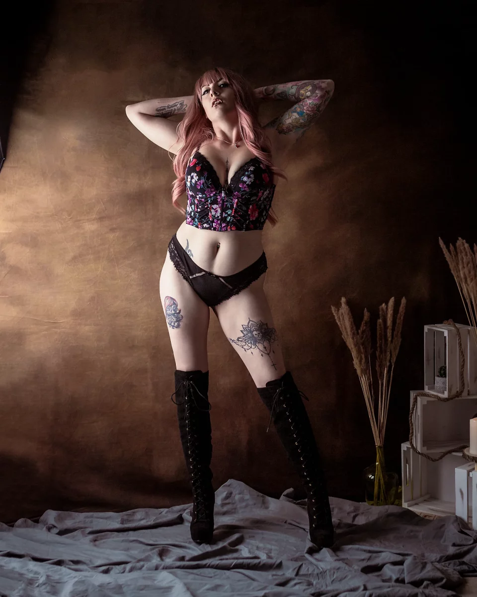 Lingerie Shooting im Studio. Dessousfotografie einer Frau in stehender Pose vor einem warm getönten, strukturierten Studiohintergrund. Die Frau steht mit leicht gespreizten Beinen, das Gewicht ungleich verteilt, wodurch eine ausgeprägte Hüftlinie entsteht. Beide Arme sind nach oben geführt und hinter dem Kopf verschränkt, wodurch sich der Oberkörper öffnet und die Brustpartie betont wird. Sie trägt eine schwarze, florale Corsage mit passendem Slip sowie hohe, geschnürte Overknee-Stiefel. Der Kopf ist leicht nach hinten geneigt, der Blick nach oben gerichtet, was eine selbstbewusste und expressive Ausstrahlung erzeugt. Tätowierungen an Armen und Beinen setzen zusätzliche visuelle Akzente und unterstützen die Linienführung des Körpers. Das Licht ist weich, aber gezielt von der Seite geführt und modelliert die Körperkonturen mit sanften Schatten, während der Hintergrund ruhig und diffus bleibt. Dekorative Elemente wie Holzkisten und Trockenpflanzen am Bildrand ergänzen die Szene subtil. Die Kombination aus offenem Posing, gezielter Lichtführung und kontrastreichem Styling erzeugt eine kraftvolle, inszenierte Bildwirkung im Bereich der Studio- und Editorial-Dessousfotografie.