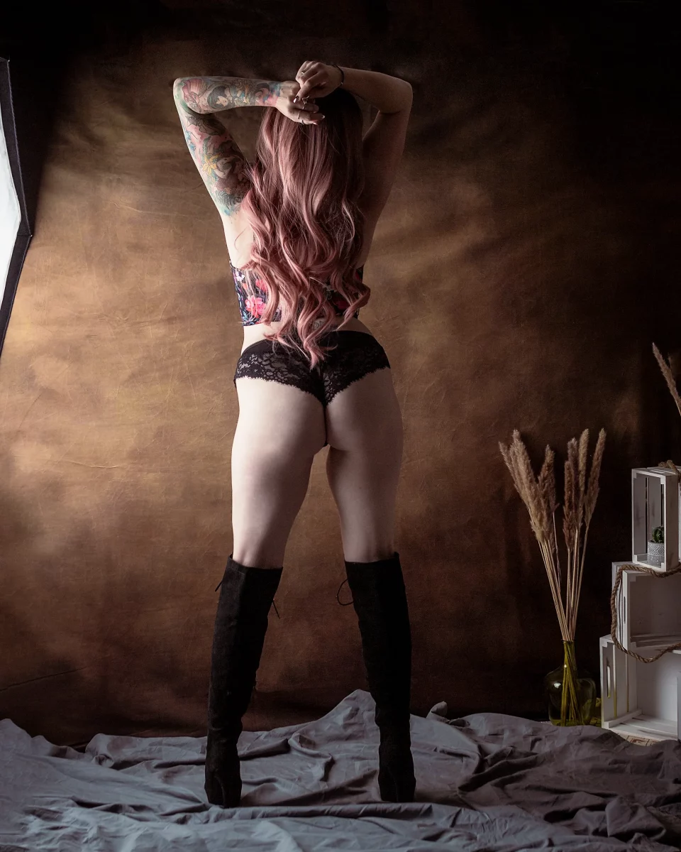 Lingerie Shooting im Studio. Dessousfotografie einer Frau in stehender Rückenansicht vor einem warm getönten, strukturierten Studiohintergrund. Die Frau steht mit leicht gespreizten Beinen, das Gewicht gleichmäßig verteilt, wodurch eine stabile, aufrechte Körperhaltung entsteht. Beide Arme sind nach oben geführt und hinter dem Kopf verschränkt, wodurch Schultern und Rücken geöffnet werden. Sie trägt eine schwarze Spitzenunterhose, eine florale Corsage und hohe, geschnürte Overknee-Stiefel. Das lange, leicht gewellte Haar fällt über den Rücken und wird vom seitlichen Licht sanft hervorgehoben. Tätowierungen an Armen ergänzen die Linienführung und setzen zusätzliche visuelle Akzente. Das Licht ist weich und seitlich geführt, modelliert die Konturen von Rücken, Hüfte und Beinen und erzeugt sanfte Schattenverläufe. Der Hintergrund bleibt ruhig und diffus, während dekorative Elemente wie Holzkisten und Trockenpflanzen am Bildrand dezent sichtbar sind. Die Kombination aus Rückenansicht, klarer Körperhaltung und gezielter Lichtführung erzeugt eine reduzierte, grafisch wirkende Bildkomposition innerhalb der Studio- und Editorial-Dessousfotografie.