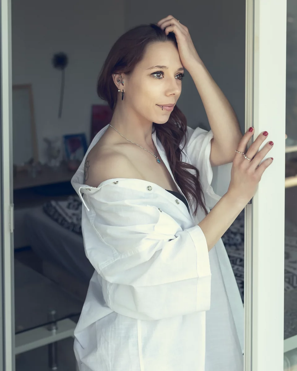 Home Shooting. Eine junge Frau steht seitlich an einer geöffneten Balkontür und hält sich leicht am Rahmen fest, während sie mit ruhigem Blick nach draußen schaut. Sie trägt ein locker sitzendes, weißes Hemd, das von der Schulter gefallen ist und eine entspannte, natürliche Wirkung erzeugt. Ihr langes Haar fällt über eine Seite, und dezenter Schmuck sowie kleine Piercings sind sichtbar. Das helle Tageslicht fällt weich von außen ein und beleuchtet ihr Gesicht sowie die feinen Strukturen des Stoffes. Der Innenraum im Hintergrund bleibt unscharf und zurückhaltend, wodurch die Szene insgesamt ruhig, intim und leicht nachdenklich wirkt.