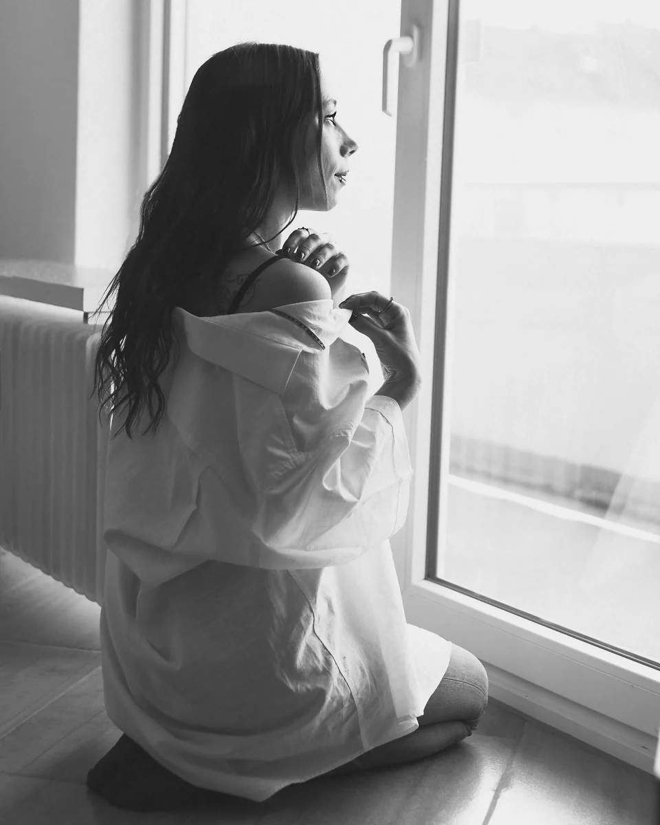Home Shooting. In einer ruhigen Schwarzweißaufnahme sitzt eine junge Frau auf dem Boden nahe einem Fenster und blickt nach draußen. Sie trägt ein locker sitzendes Hemd, das von der Schulter gefallen ist, und hält es leicht mit einer Hand fest. Ihr langes Haar fällt über den Rücken, während das weiche Tageslicht von außen ihr Profil sanft beleuchtet und feine Kontraste erzeugt. Der Innenraum bleibt schlicht und zurückhaltend, wodurch der Fokus auf der stillen, nachdenklichen Stimmung und der ruhigen Körperhaltung liegt.