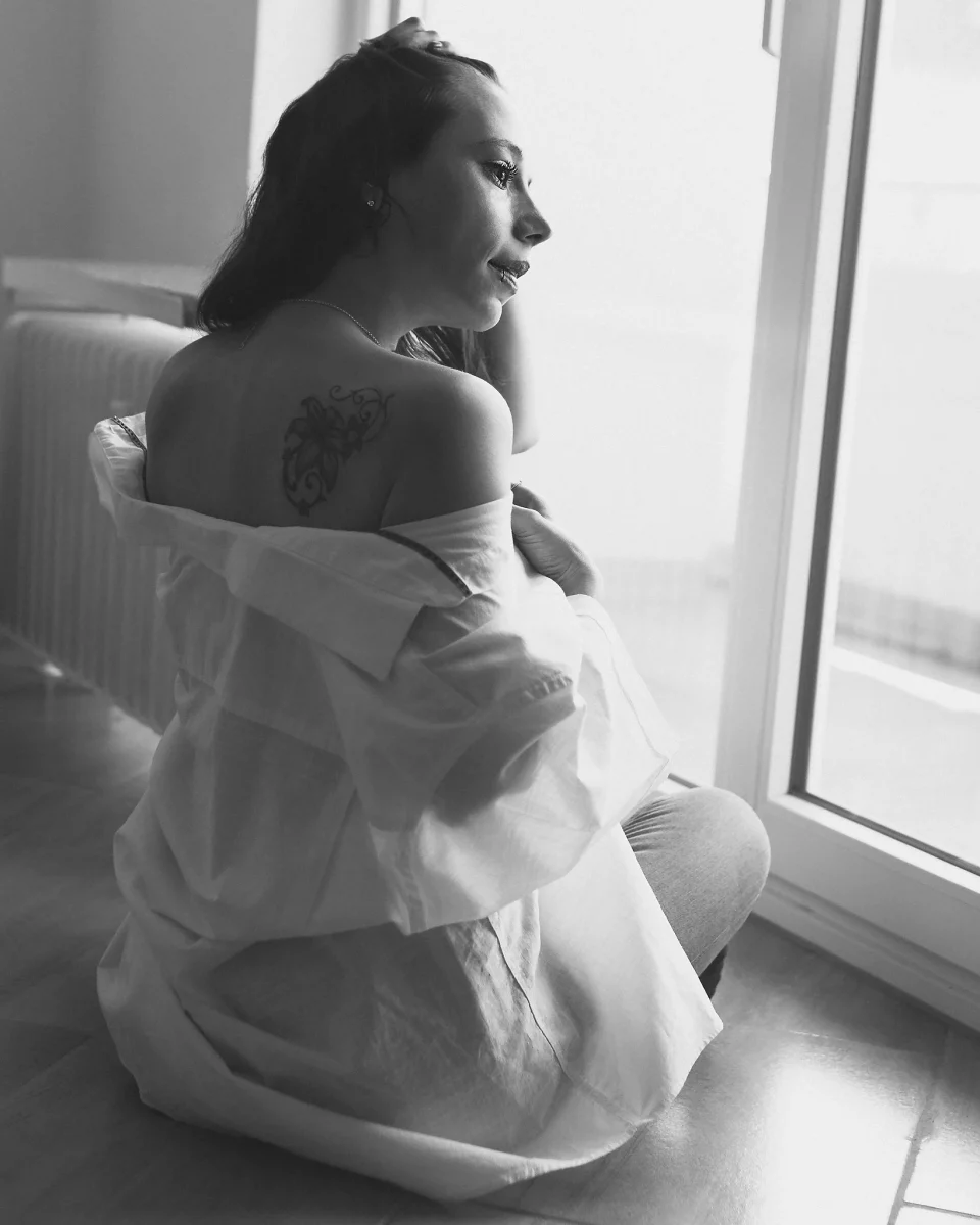 Home Shooting. In einer ruhigen Schwarzweißszene sitzt eine junge Frau auf dem Boden nahe einem Fenster und wendet ihren Blick nach draußen. Ihr locker getragenes Hemd ist von der Schulter gerutscht und gibt den Blick auf ein Tattoo frei, während sie den Stoff leicht mit der Hand hält. Das seitlich einfallende Tageslicht modelliert sanft ihr Gesicht und ihren Rücken und erzeugt weiche Kontraste. Der Raum bleibt schlicht und zurückhaltend, wodurch die Szene eine ruhige, intime und leicht nachdenkliche Atmosphäre erhält.
