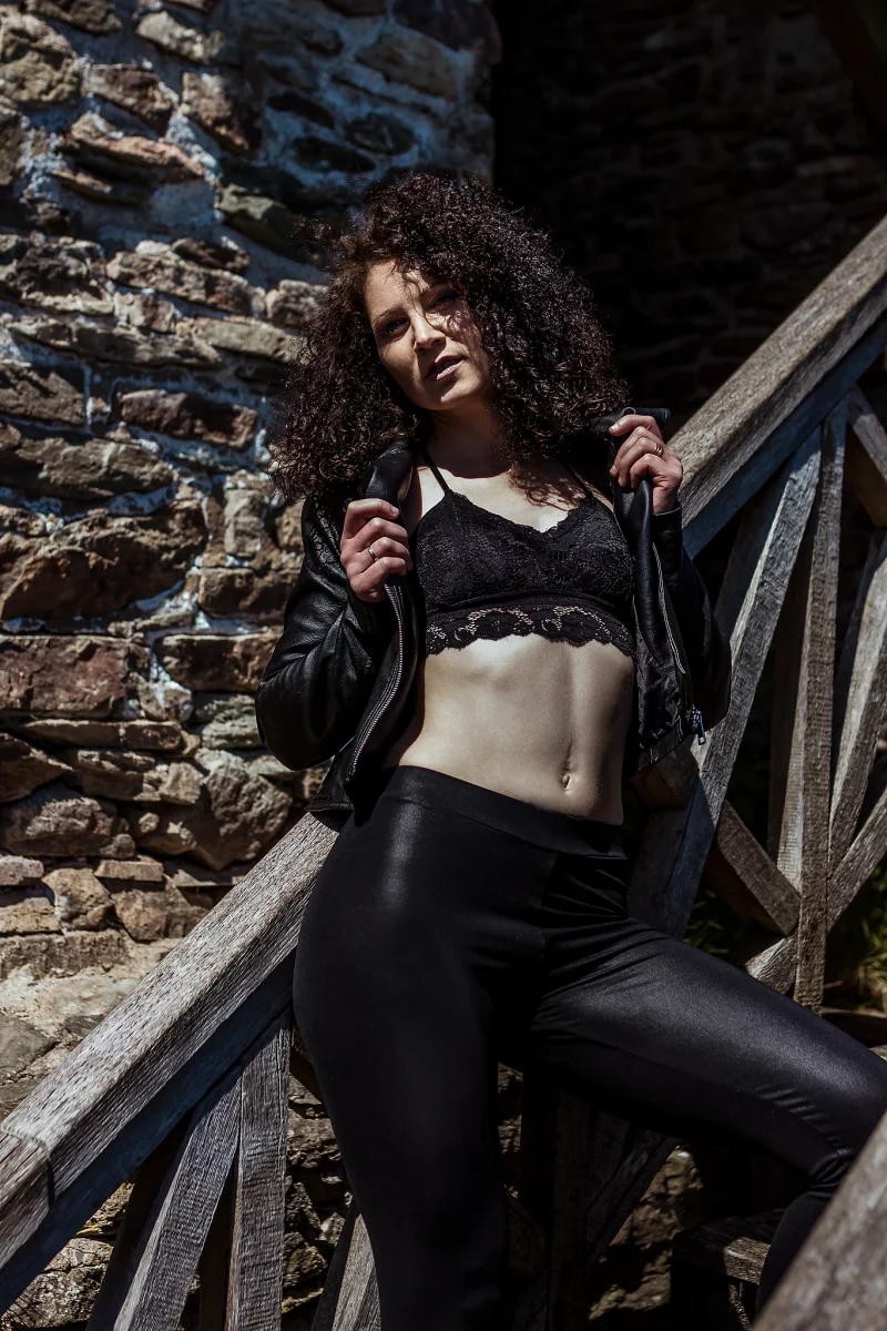 Lingerie Shooting auf der Grimburg. Dessousfotografie einer Frau in einem Außenbereich mit rustikaler Steinmauer und hölzerner Treppe. Die Frau steht auf einer Treppenstufe, der Körper leicht nach hinten an das Geländer gelehnt, ein Bein erhöht positioniert. Sie trägt ein schwarzes Spitzen-Bralette, eine enge, glänzende Hose und eine offene Lederjacke, die mit beiden Händen leicht gehalten wird. Der Oberkörper ist leicht nach hinten geneigt, wodurch die Bauchpartie betont wird. Der Blick ist zur Kamera gerichtet, mit leicht geneigtem Kopf und einer ruhigen, selbstbewussten Ausstrahlung. Das Licht ist hartes Tageslicht mit starken Kontrasten und klaren Schatten, wodurch Hautstruktur, Stoffe und die raue Oberfläche der Steine deutlich hervorgehoben werden. Die Kombination aus natürlicher Umgebung, kantiger Architektur und kontrastreichem Licht erzeugt eine kraftvolle, leicht urbane Bildwirkung im Bereich der Outdoor- und Editorial-Dessousfotografie.