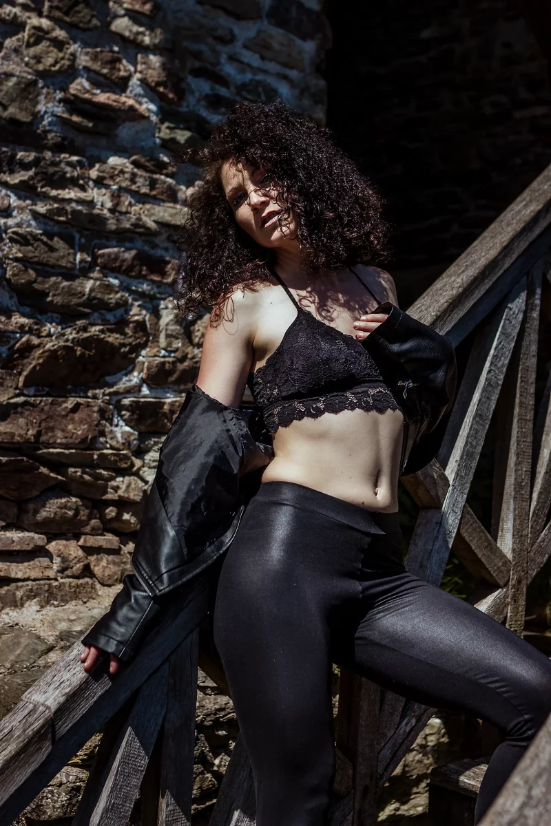 Lingerie Shooting auf der Grimburg. Dessousfotografie einer Frau in einem Außenbereich mit rauer Steinmauer und hölzerner Treppe. Die Frau lehnt seitlich an einem Holzgeländer, ein Bein erhöht auf der Treppe positioniert, wodurch eine diagonale Körperhaltung entsteht. Sie trägt ein schwarzes Spitzen-Bralette und eine eng anliegende, glänzende Hose, während eine Lederjacke locker von den Schultern rutscht. Eine Hand hält die Jacke leicht am Körper, die andere liegt entspannt am Geländer. Der Oberkörper ist leicht gedreht, der Kopf zur Seite geneigt und der Blick in Richtung Kamera gerichtet. Das harte Tageslicht erzeugt starke Kontraste und betont die Strukturen von Haut, Stoff und Steinoberfläche. Die Kombination aus natürlicher Umgebung, groben Materialien und kontrastreicher Lichtführung verstärkt die körperliche Präsenz und erzeugt eine kraftvolle, leicht raue Bildwirkung im Bereich der Outdoor- und Editorial-Dessousfotografie.