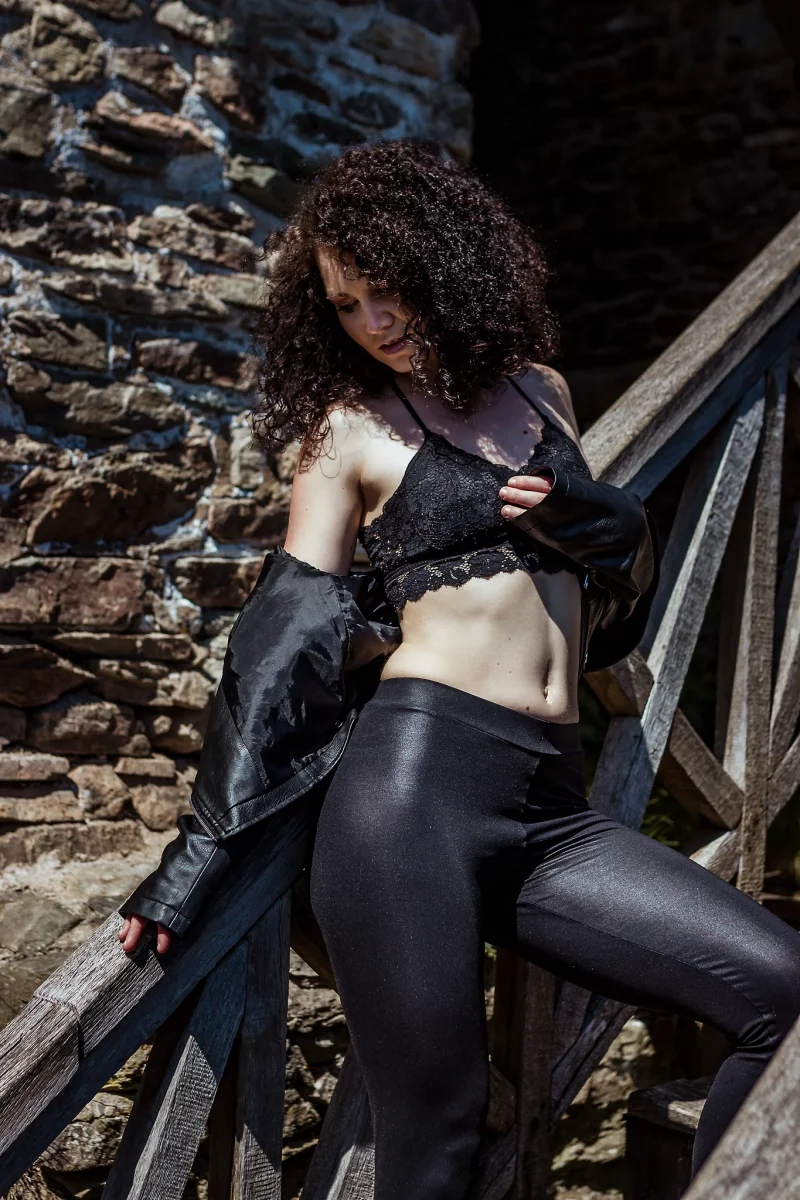 Lingerie Shooting auf der Grimburg. Dessousfotografie einer Frau auf einer hölzernen Treppe vor einer rauen Steinmauer im Außenbereich. Die Frau lehnt seitlich am Geländer, ein Bein auf einer höheren Stufe positioniert, wodurch eine schräge, dynamische Körperlinie entsteht. Sie trägt ein schwarzes Spitzen-Bralette und eine eng anliegende, glänzende Hose, während eine Lederjacke locker von den Schultern rutscht. Eine Hand hält die Jacke leicht am Körper, die andere liegt am Geländer. Der Kopf ist nach unten geneigt, der Blick auf den eigenen Oberkörper gerichtet, wodurch eine zurückgenommene, ruhige Bildwirkung entsteht. Das harte Sonnenlicht erzeugt starke Kontraste und hebt die Struktur von Haut, Stoffen und der Steinoberfläche deutlich hervor. Die Kombination aus diagonaler Körperhaltung, rauer Architektur und prägnanter Lichtführung erzeugt eine kraftvolle, zugleich reduzierte Bildkomposition im Bereich der Outdoor- und Editorial-Dessousfotografie.