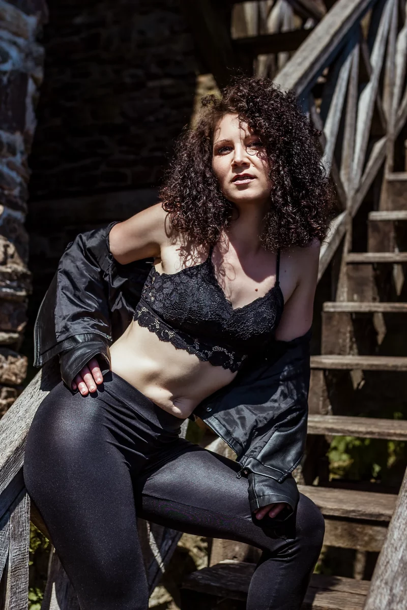 Lingerie Shooting auf der Grimburg. Dessousfotografie einer Frau auf einer hölzernen Außentreppe vor einer grob strukturierten Steinwand. Die Frau sitzt seitlich auf dem Geländer, ein Bein angewinkelt auf der Stufe, das andere tiefer positioniert, wodurch eine kompakte, stabile Körperhaltung entsteht. Sie trägt ein schwarzes Spitzen-Bralette und eine eng anliegende, glänzende Hose. Eine schwarze Lederjacke ist locker über die Arme gelegt und teilweise von den Schultern gerutscht. Der Oberkörper ist leicht nach vorne geneigt, wodurch die Bauch- und Rumpfpartie betont wird. Eine Hand liegt auf dem Oberschenkel, die andere stützt sich am Geländer ab. Der Blick ist direkt zur Kamera gerichtet, mit leicht angehobenem Kinn und einer ruhigen, selbstbewussten Ausstrahlung. Das harte Sonnenlicht erzeugt ausgeprägte Licht- und Schattenkanten und hebt die Struktur von Haut, Stoff und Holz deutlich hervor. Die Kombination aus körpernaher Perspektive, klarer Pose und rauer Umgebung erzeugt eine kraftvolle, präsente Bildwirkung im Bereich der Outdoor- und Editorial-Dessousfotografie.