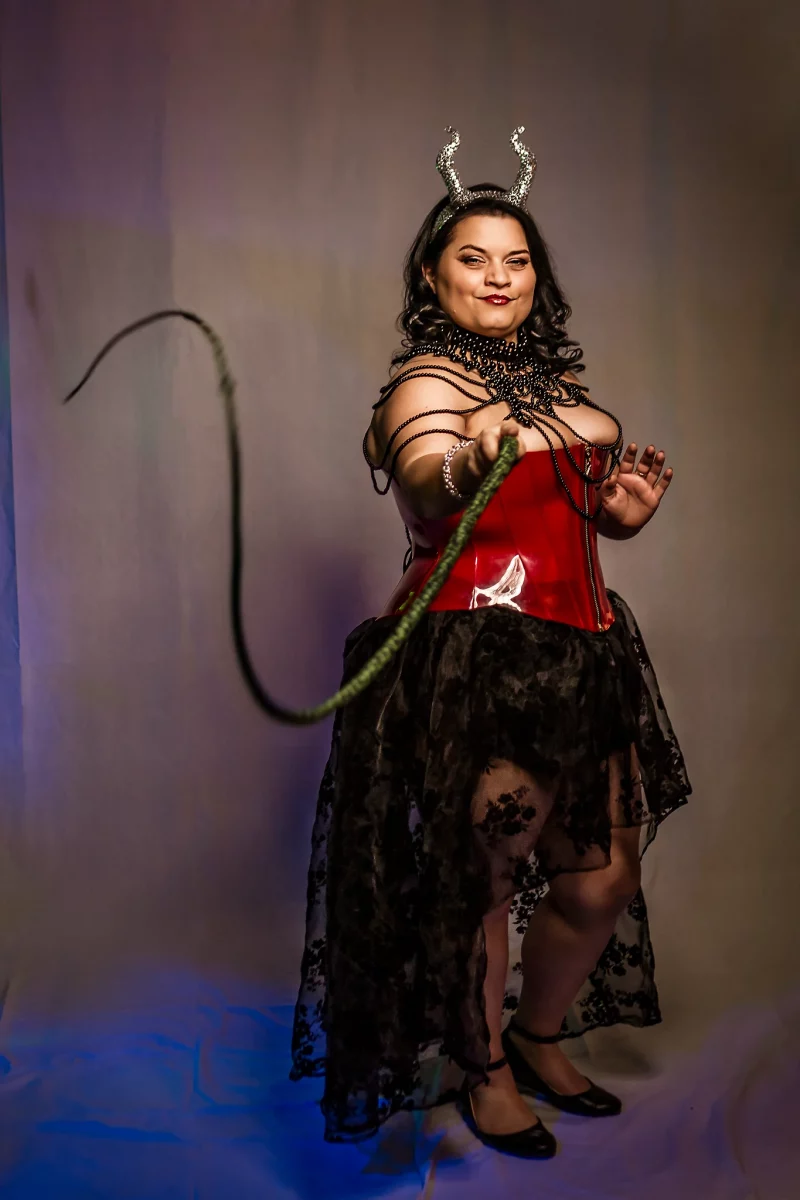 Lingerie Shooting mit Miss Saphir. Inszenierte Dessous- und Fetischfotografie im Studio vor einem neutralen, weich verlaufenden Hintergrund. Eine Frau steht frontal zur Kamera, der Körper leicht in eine diagonale Haltung gebracht, ein Bein minimal nach vorne gesetzt. Sie trägt ein rotes, glänzendes Korsett, einen schwarzen, transparenten Spitzenrock sowie schwarze High Heels. Um den Hals und die Schultern legt sich ein dekoratives, kettenartiges Accessoire, ergänzt durch ein Haar-Accessoire in Form von Hörnern. In einer Hand hält sie eine Peitsche, die in einer geschwungenen Linie durch den Vordergrund verläuft und Dynamik in die Bildkomposition bringt. Der Blick ist direkt zur Kamera gerichtet, mit leichtem Lächeln und selbstbewusster Ausstrahlung. Das Licht ist weich geführt mit sanften Farbakzenten im Hintergrund, wodurch Hauttöne und Materialien wie Lack, Spitze und Metall differenziert herausgearbeitet werden. Die Kombination aus Pose, Accessoires und Lichtgestaltung erzeugt eine klare, inszenierte Bildwirkung im Bereich der Studio- und Fetisch-Dessousfotografie.