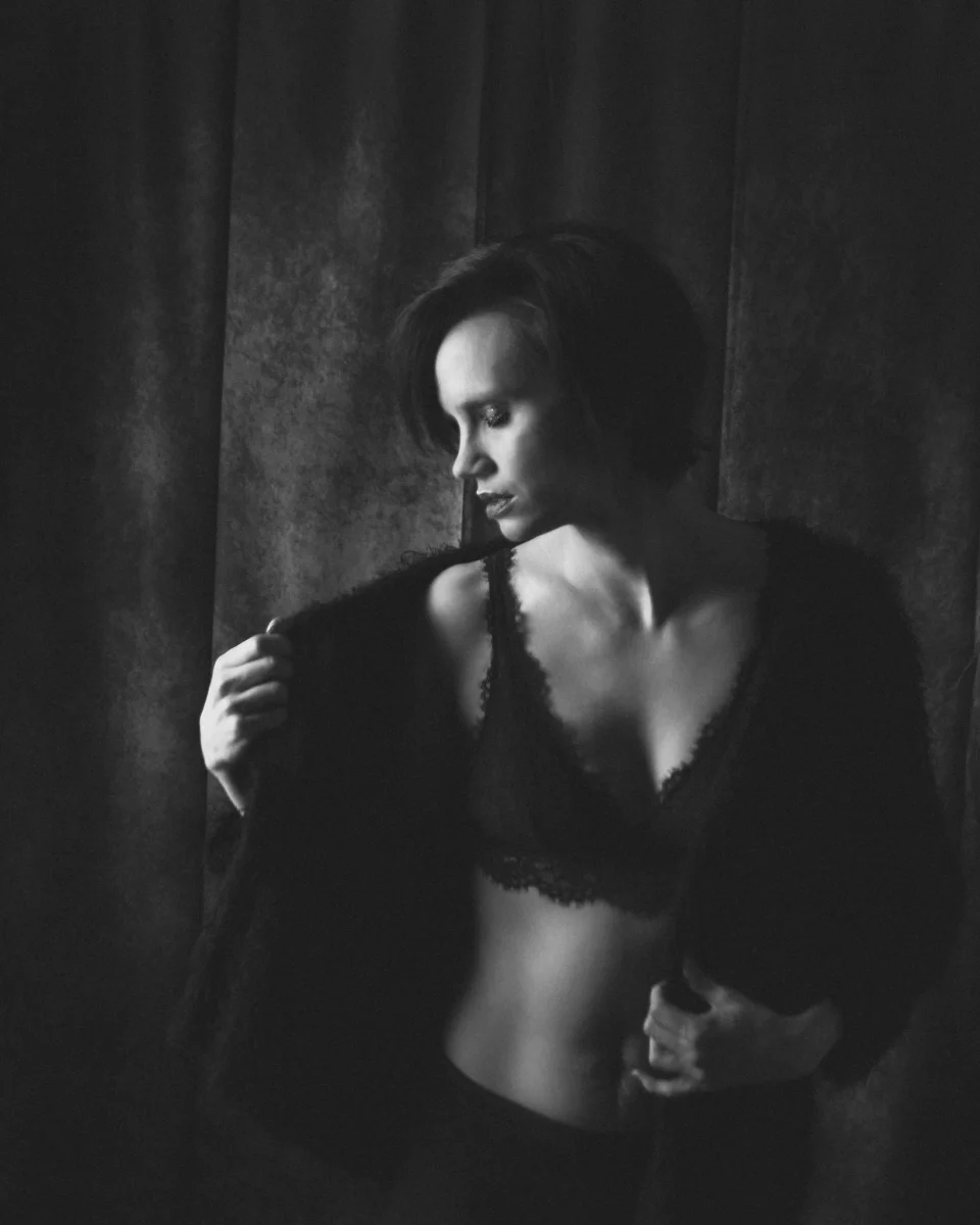 Lingerie Shooting in Schwarzweiß im Hochformat. Schwarz-weiße Dessousfotografie einer Frau vor einem dunklen, strukturierten Vorhang im Studio. Die Frau steht leicht seitlich zur Kamera, der Oberkörper sanft gedreht. Sie trägt ein dunkles Spitzen-Bralette sowie eine offene, weiche Jackel, die sie mit beiden Händen leicht vom Körper wegzieht. Der Kopf ist nach unten geneigt, der Blick auf die eigene Schulter gerichtet, wodurch eine ruhige, introspektive Bildwirkung entsteht. Das Licht ist weich und gerichtet, seitlich einfallend, und modelliert Gesicht, Schlüsselbein und Oberkörper mit sanften Übergängen. Die Tonwertverteilung ist kontrastreich, mit tiefen Schatten und hellen Hautpartien, wodurch die Struktur von Spitze und Stoff sowie die Konturen des Körpers betont werden. Die reduzierte Farbigkeit verstärkt die grafische Wirkung und lenkt den Fokus auf Form, Licht und Textur im Bereich der klassischen Schwarz-Weiß-Dessousfotografie.