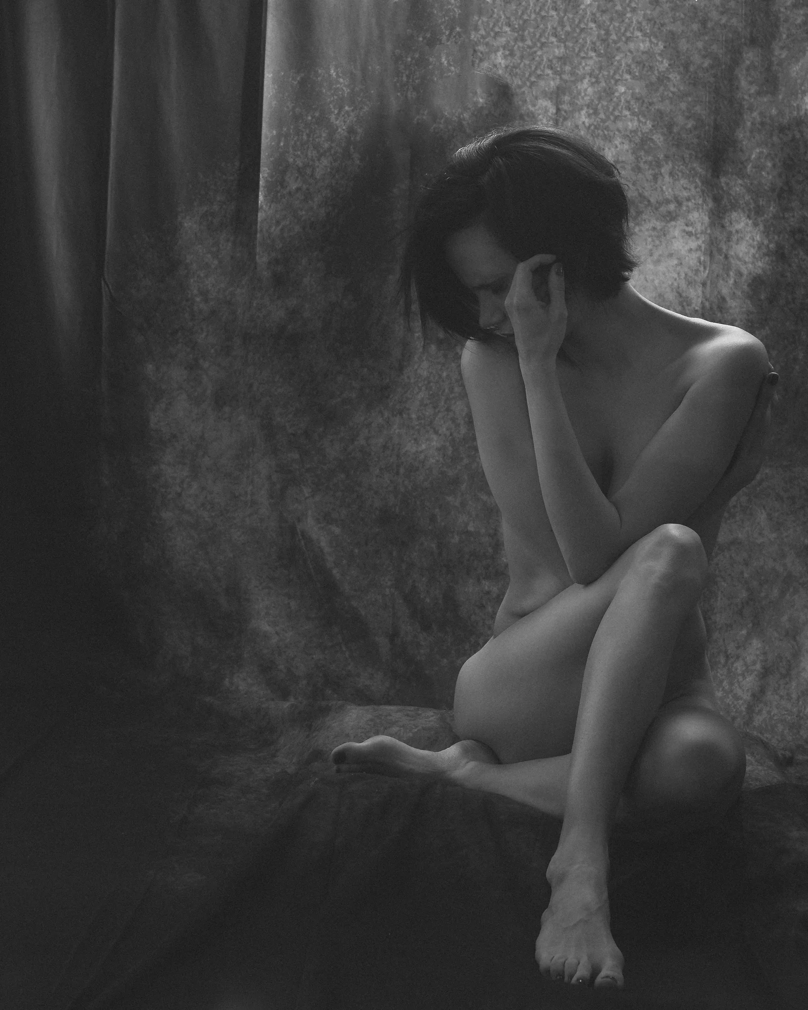 Nude Shoot mit Sarah in Schwarzweiß. Schwarzweiß-Aktfotografie einer Frau, die auf einem Podest vor einem strukturierten Stoffhintergrund sitzt. Ihr Körper ist leicht eingedreht, die Beine angewinkelt, während ein Arm schützend über den Oberkörper gelegt ist. Mit der anderen Hand berührt sie ihr Gesicht, der Blick ist gesenkt und nach innen gerichtet. Das weiche, gerichtete Licht modelliert sanft die Konturen ihres Körpers und erzeugt feine Übergänge zwischen Licht und Schatten. Die reduzierte Bildsprache und die ruhige, geschlossene Körperhaltung vermitteln eine intime, nachdenkliche und beinahe fragile Stimmung im Sinne klassischer Fine-Art-Aktfotografie.