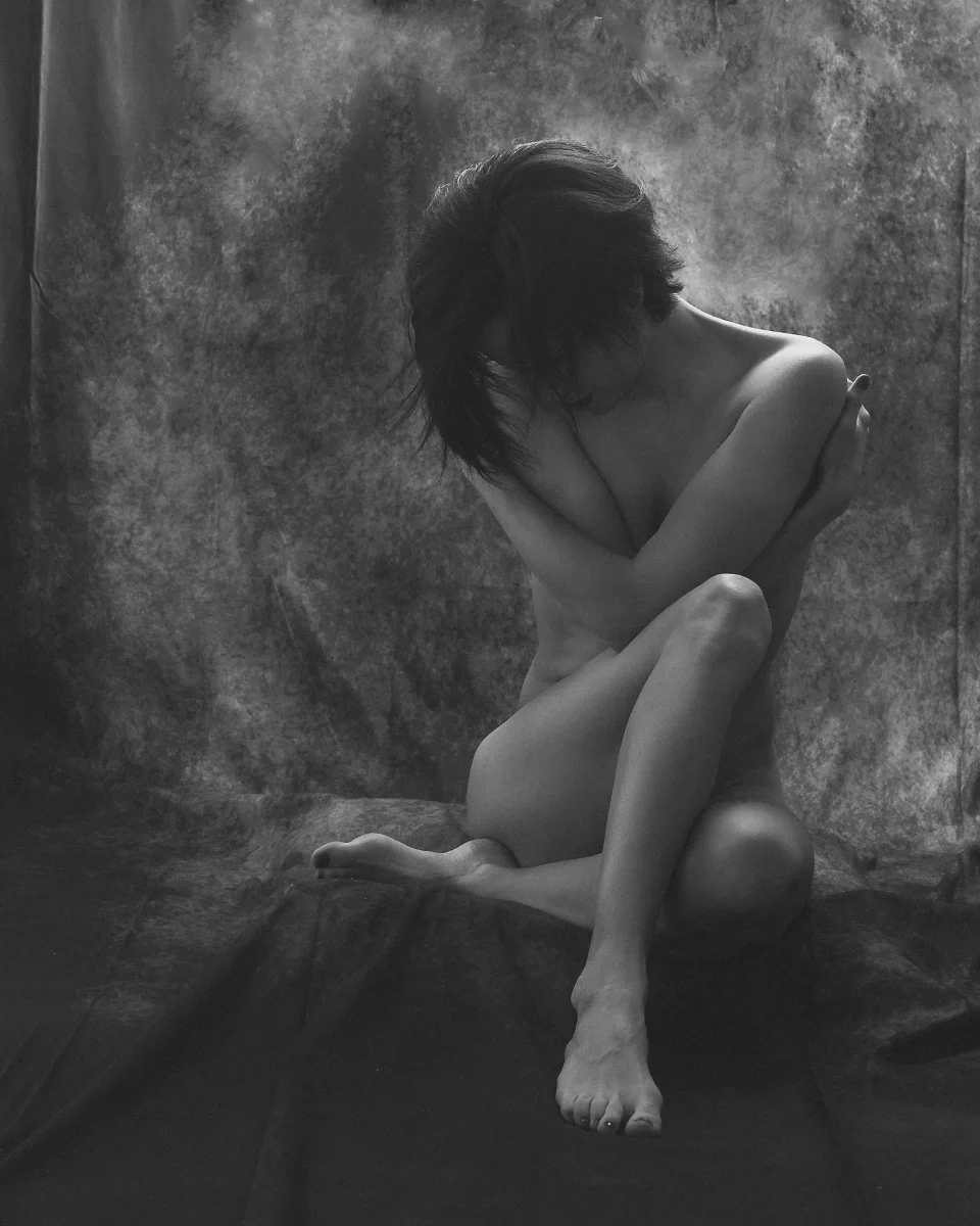Nude Shoot in Schwarzweiß.Schwarzweiß-Aktfotografie einer sitzenden Frau vor einem weich strukturierten Stoffhintergrund. Ihr Körper ist kompakt und in sich geschlossen positioniert, die Beine angewinkelt und übereinandergeschlagen, während die Arme schützend über den Oberkörper gelegt sind. Der Kopf ist nach vorne geneigt, das Gesicht teilweise vom Haar verdeckt, wodurch der Blick verborgen bleibt. Das weiche, diffuse Licht umspielt die Konturen von Schultern, Beinen und Füßen und erzeugt sanfte Übergänge zwischen Licht und Schatten. Die reduzierte Komposition, die geschlossene Körperhaltung und die gedämpfte Tonalität verleihen der Aufnahme eine ruhige, intime und leicht verletzliche Stimmung im klassischen Kontext der Fine-Art-Aktfotografie.