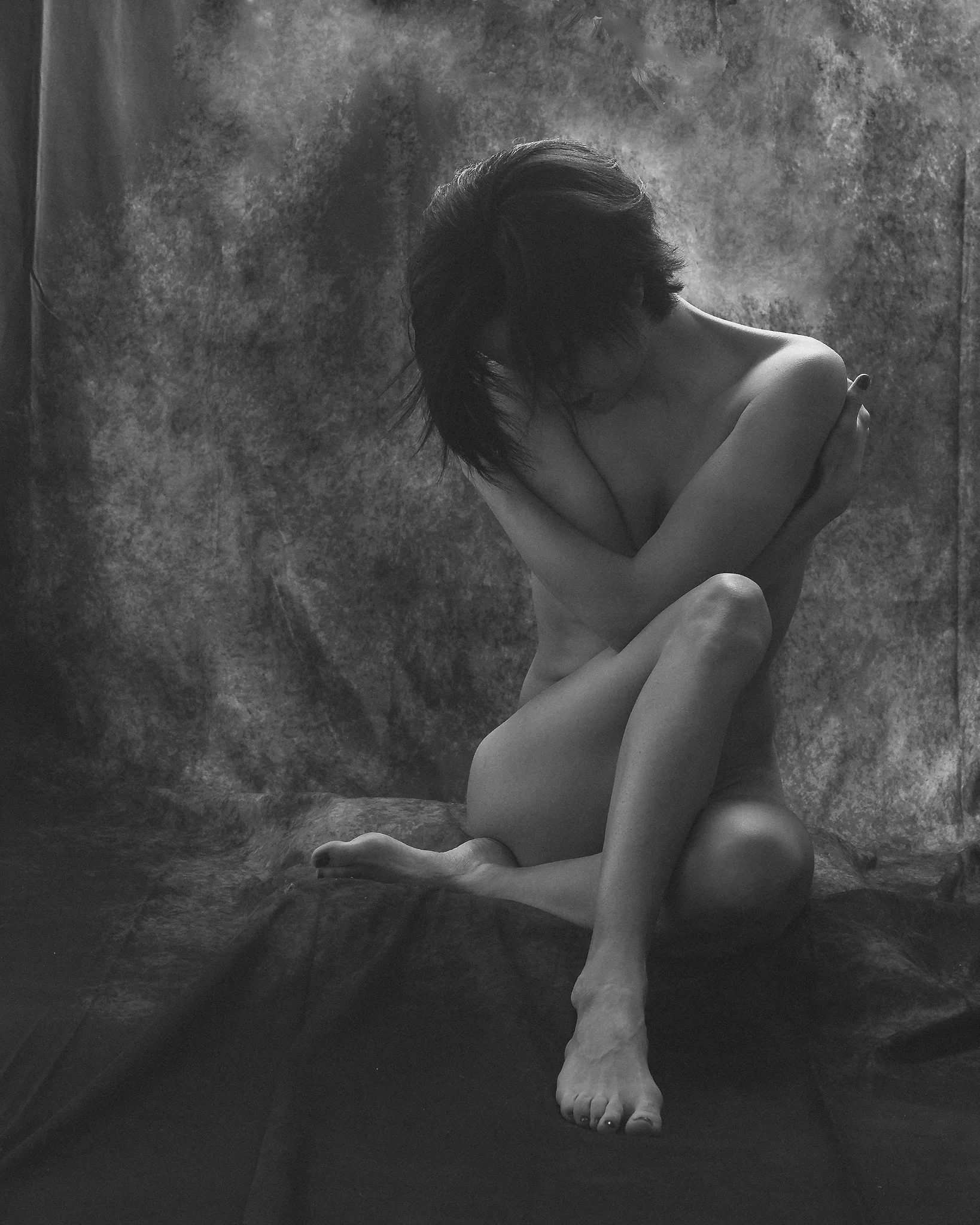 Nude Shoot in Schwarzweiß.Schwarzweiß-Aktfotografie einer sitzenden Frau vor einem weich strukturierten Stoffhintergrund. Ihr Körper ist kompakt und in sich geschlossen positioniert, die Beine angewinkelt und übereinandergeschlagen, während die Arme schützend über den Oberkörper gelegt sind. Der Kopf ist nach vorne geneigt, das Gesicht teilweise vom Haar verdeckt, wodurch der Blick verborgen bleibt. Das weiche, diffuse Licht umspielt die Konturen von Schultern, Beinen und Füßen und erzeugt sanfte Übergänge zwischen Licht und Schatten. Die reduzierte Komposition, die geschlossene Körperhaltung und die gedämpfte Tonalität verleihen der Aufnahme eine ruhige, intime und leicht verletzliche Stimmung im klassischen Kontext der Fine-Art-Aktfotografie.