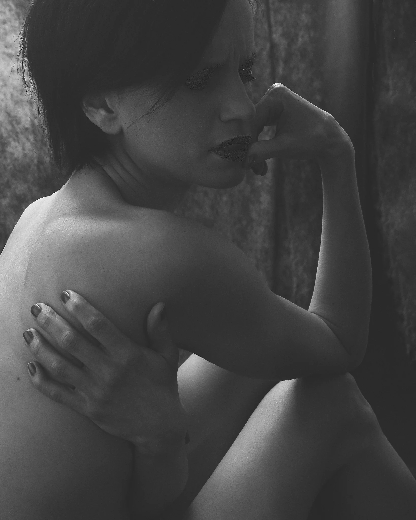 @mrs._sarah_w Home Shooting. In einer kontrastreichen Schwarzweißaufnahme sitzt eine junge Frau in einer geschlossenen, nach innen gerichteten Haltung. Sie umfasst mit einer Hand ihre Schulter, während die andere Hand nahe ihrem Gesicht liegt. Ihr Blick ist nach unten gerichtet, wodurch eine ruhige, introspektive Stimmung entsteht. Das Licht fällt weich von der Seite ein und modelliert sanft die Konturen ihres Gesichts und Körpers, während der Hintergrund dunkel und zurückhaltend bleibt. Die reduzierte Bildsprache verstärkt die intime, ruhige Wirkung der Szene und lenkt den Fokus vollständig auf Ausdruck und Haltung.