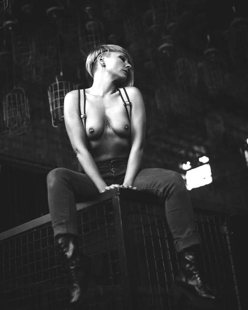 @totalblue_model Lost Places - Schwarzweißes Aktporträt einer Frau mit kurzen blonden Haaren, die auf einer erhöhten Metallkonstruktion sitzt; sie trägt Hose, Hosenträger und Stiefel, ist oberkörperfrei und blickt seitlich aus dem Bild, während das harte, gerichtete Licht starke Kontraste erzeugt und die industrielle Umgebung mit hängenden Drahtkörben eine raue, grafische Bildwirkung unterstützt.