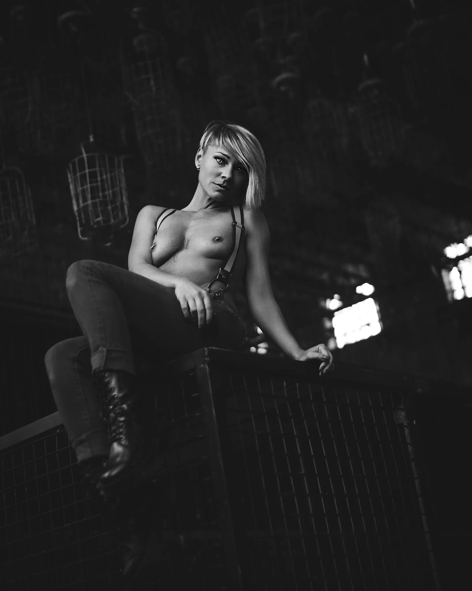 Lost Places - Schwarzweißes Aktporträt einer Frau mit kurzen blonden Haaren, die lässig auf einer erhöhten Metallkonstruktion sitzt; sie trägt Hose, Hosenträger und schwere Stiefel, ist oberkörperfrei und blickt direkt zur Kamera, während hartes, gerichtetes Licht ihre Körperformen betont und die industrielle Umgebung mit hängenden Drahtkörben eine düstere, kontrastreiche Atmosphäre schafft.