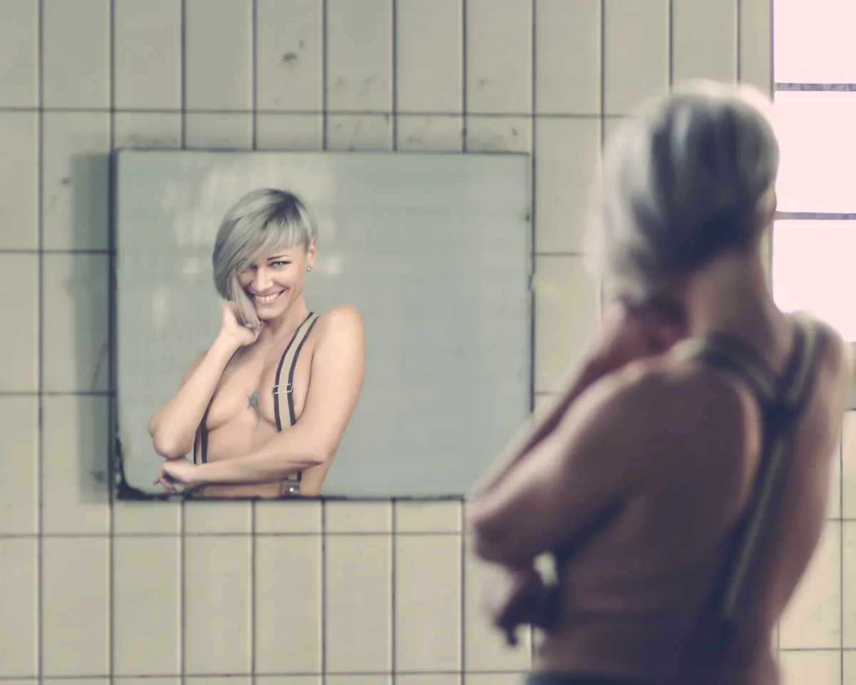 @totalblue_model Lost Places - Teilakt-Porträt einer Frau mit kurzem, hellem Haar vor einem Wandspiegel in einem gekachelten Raum; ihr lächelndes Spiegelbild steht im Fokus, während ihr Körper im Vordergrund weich unscharf bleibt und das diffuse, helle Licht eine ruhige, intime Atmosphäre erzeugt.