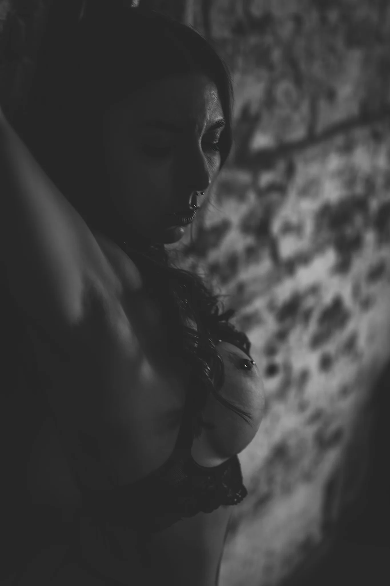 Studio - Schwarzweißes Detailporträt einer tätowierten Person in seitlicher Ansicht, mit nach unten geneigtem Blick und erhobenen Armen; der Oberkörper ist nur teilweise von einem offenen Dessous bedeckt, wodurch Licht und Schatten die Formen und Hautstruktur betonen. Das harte, gerichtete Licht erzeugt starke Kontraste und hebt Gesichtszüge sowie Konturen hervor, während die raue Backsteinwand im Hintergrund weich ausläuft und die reduzierte, intime Bildwirkung unterstützt.