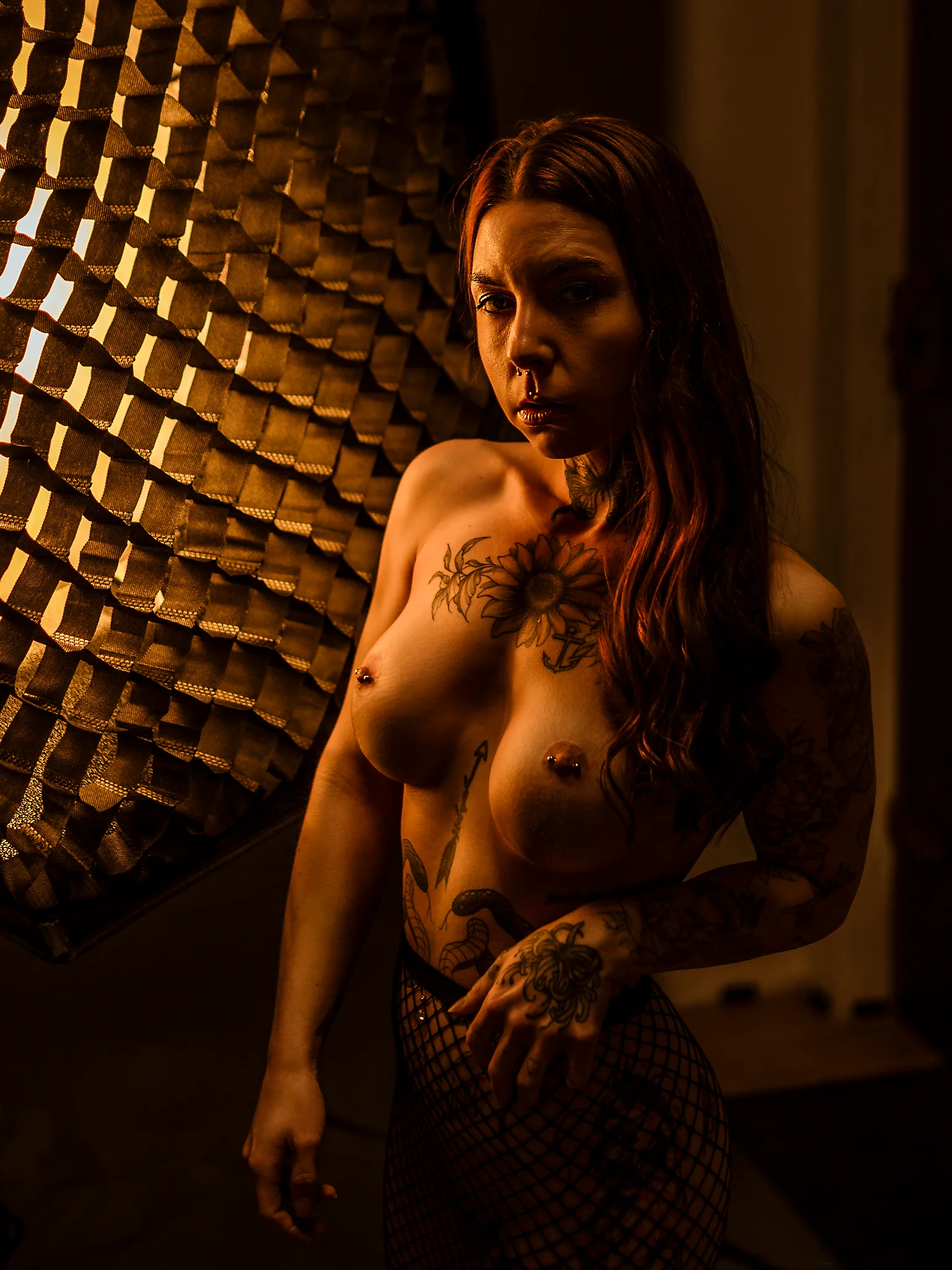 Studio - Sinnliches Low-Key-Porträt einer tätowierten Frau in warmem, amberfarbenem Licht, seitlich von einer großen Softbox beleuchtet; weiche Schatten modellieren Gesicht und Oberkörper, während der dunkle Hintergrund und das gerichtete Licht eine intime, filmische Atmosphäre erzeugen.