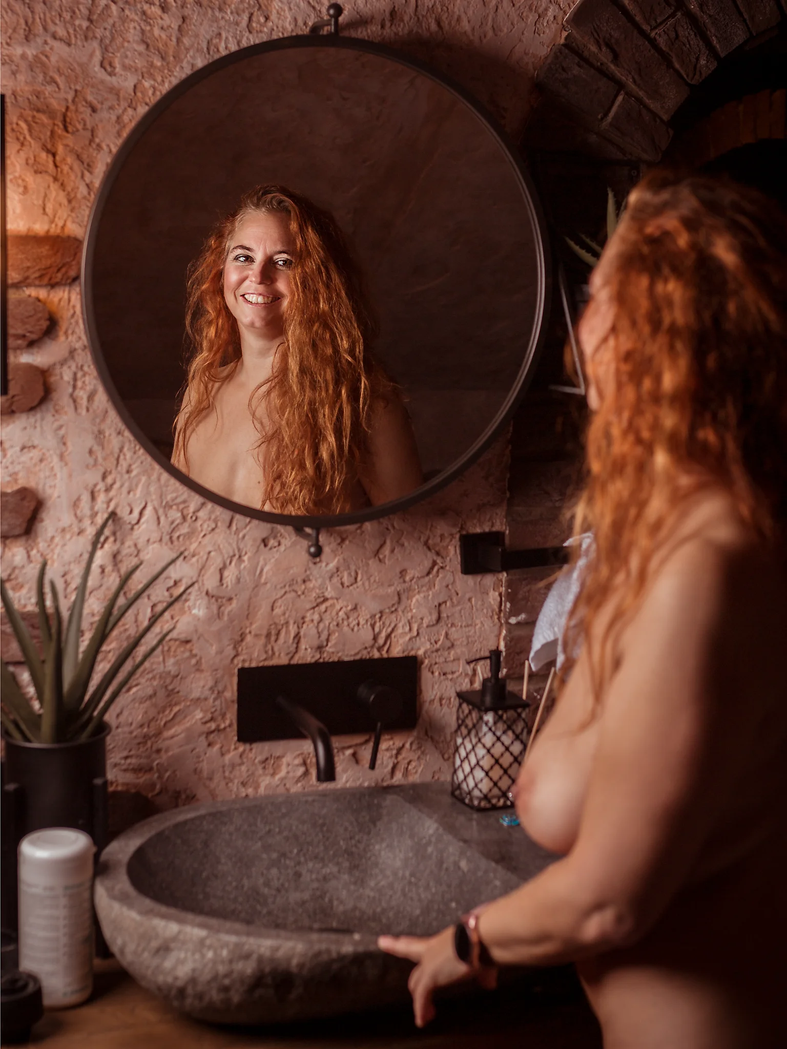 Nude Shooting im Sweet Poison mit Nicole. Aktfotografie einer Frau vor einem Waschbecken, deren Gesicht im runden Wandspiegel sichtbar wird. Während ihr Körper im Vordergrund leicht unscharf bleibt, zeigt die Spiegelung ein offenes, natürliches Lächeln und lenkt den Blick des Betrachters gezielt auf den Ausdruck. Die warme, weiche Lichtstimmung betont die natürlichen Hauttöne sowie die Struktur der Wand und schafft eine intime, wohnliche Atmosphäre. Elemente wie das Steinwaschbecken, Pflanzen und dezente Accessoires rahmen die Szene und verleihen ihr eine authentische, ruhige Alltagsnähe. Die bewusste Trennung von direkter Ansicht und Spiegelbild erzeugt eine subtile Spannung zwischen Wahrnehmung und Reflexion im Kontext der Fine-Art-Aktfotografie.