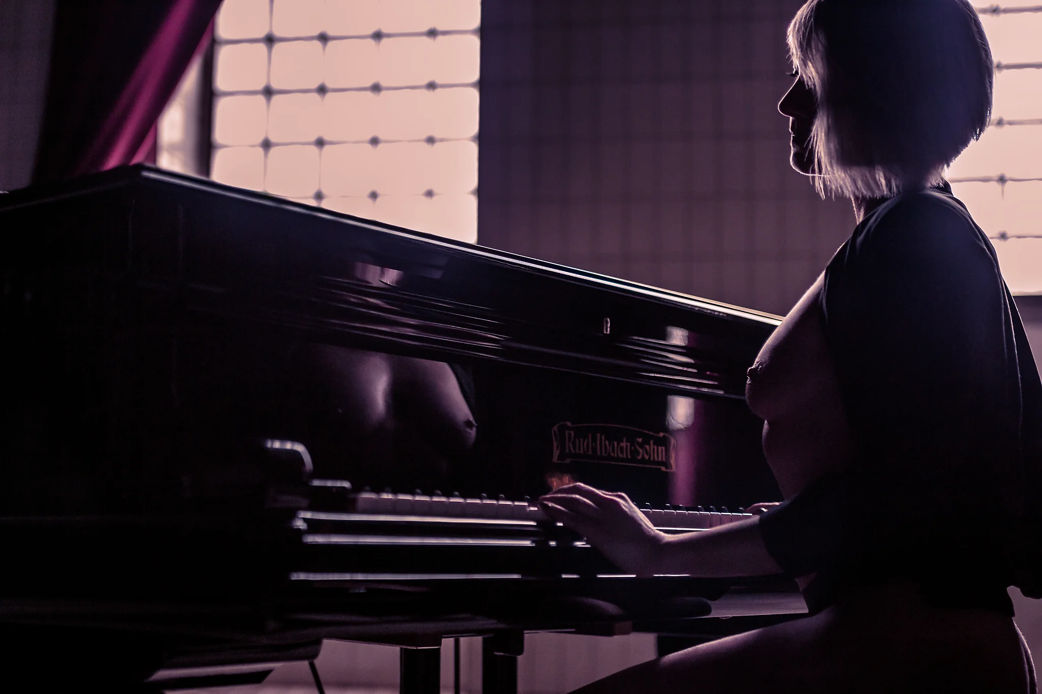 Nude Aktfotografie einer Frau, die seitlich an einem Klavier sitzt, inszeniert in einem Raum mit großem Fenster im Hintergrund. Ihr Körper ist im Profil zu sehen, die Hände ruhen auf den Tasten, während sie ruhig und konzentriert in die Szene hineinblickt. Ein offenes Kleidungsstück umrahmt den Oberkörper und schafft einen spannungsvollen Kontrast zwischen Stoff und nackter Haut. Das Licht fällt von hinten durch das Fenster und zeichnet eine klare Silhouette, während warme, leicht violette Farbtöne die Schattenbereiche durchziehen. Die glänzende Oberfläche des Klaviers reflektiert Teile des Körpers und verstärkt die visuelle Tiefe. Die Aufnahme verbindet klassisch