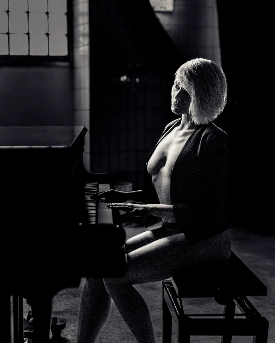 Nude in b&w at the Piano. Schwarzweiß-Aktfotografie einer Frau, die seitlich an einem Klavier sitzt. Ihr Körper ist aufrecht positioniert, die Hände ruhen leicht auf den Tasten, während sie den Blick ruhig und direkt zur Kamera richtet. Ein offenes Jackett umrahmt den Oberkörper und setzt einen subtilen Kontrast zwischen Bekleidung und nackter Haut. Das gerichtete Licht fällt seitlich ein und modelliert Gesicht, Schultern und Körper in klaren Hell-Dunkel-Kontrasten, wodurch eine starke plastische Wirkung entsteht. Der Raum im Hintergrund bleibt reduziert und dunkel, wodurch der Fokus vollständig auf der Figur und der Interaktion mit dem Instrument liegt. Die Aufnahme verbindet klassische Aktfotografie mit einer ruhigen, fast filmischen Inszenierung.