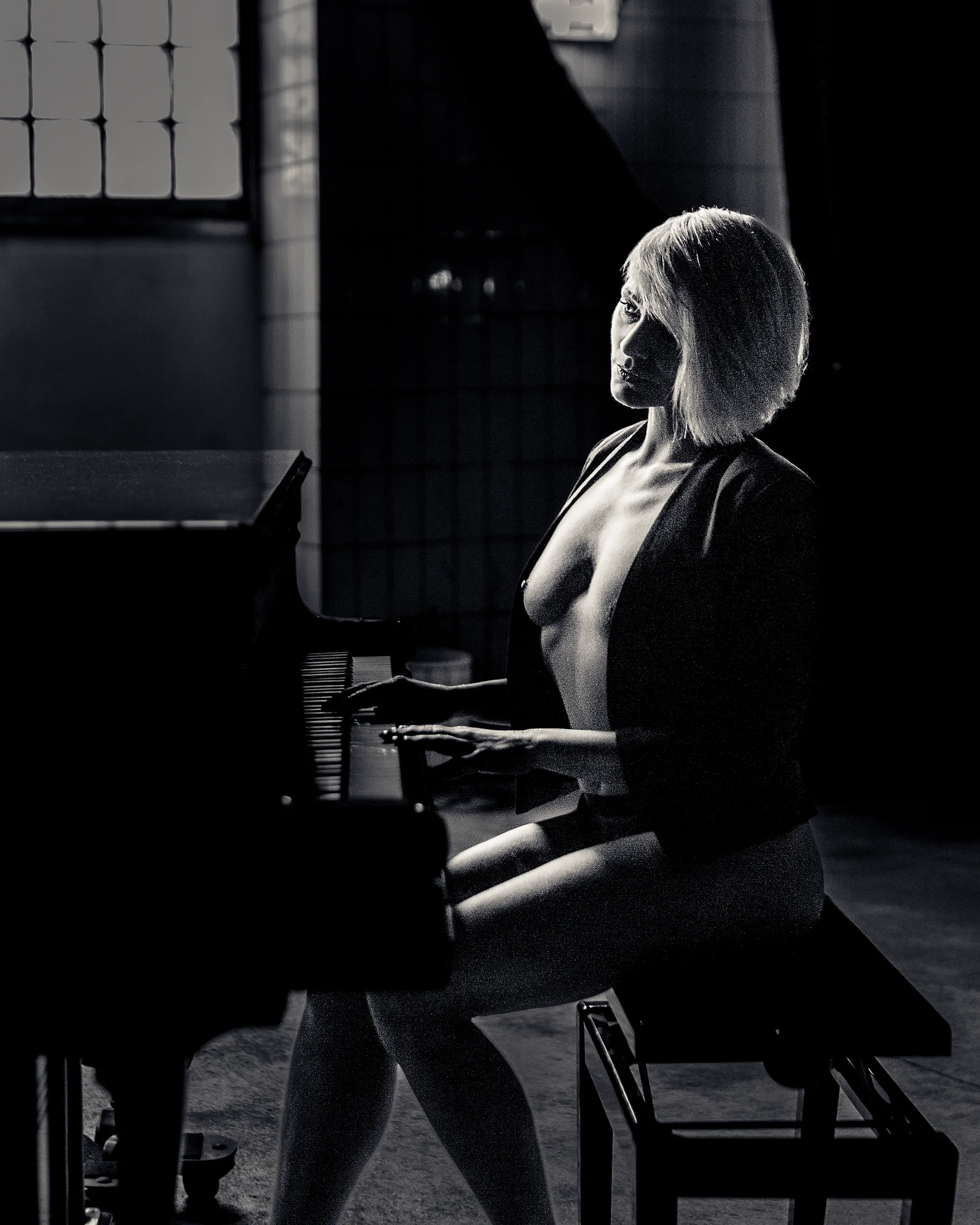 Nude in b&w at the Piano. Schwarzweiß-Aktfotografie einer Frau, die seitlich an einem Klavier sitzt. Ihr Körper ist aufrecht positioniert, die Hände ruhen leicht auf den Tasten, während sie den Blick ruhig und direkt zur Kamera richtet. Ein offenes Jackett umrahmt den Oberkörper und setzt einen subtilen Kontrast zwischen Bekleidung und nackter Haut. Das gerichtete Licht fällt seitlich ein und modelliert Gesicht, Schultern und Körper in klaren Hell-Dunkel-Kontrasten, wodurch eine starke plastische Wirkung entsteht. Der Raum im Hintergrund bleibt reduziert und dunkel, wodurch der Fokus vollständig auf der Figur und der Interaktion mit dem Instrument liegt. Die Aufnahme verbindet klassische Aktfotografie mit einer ruhigen, fast filmischen Inszenierung.
