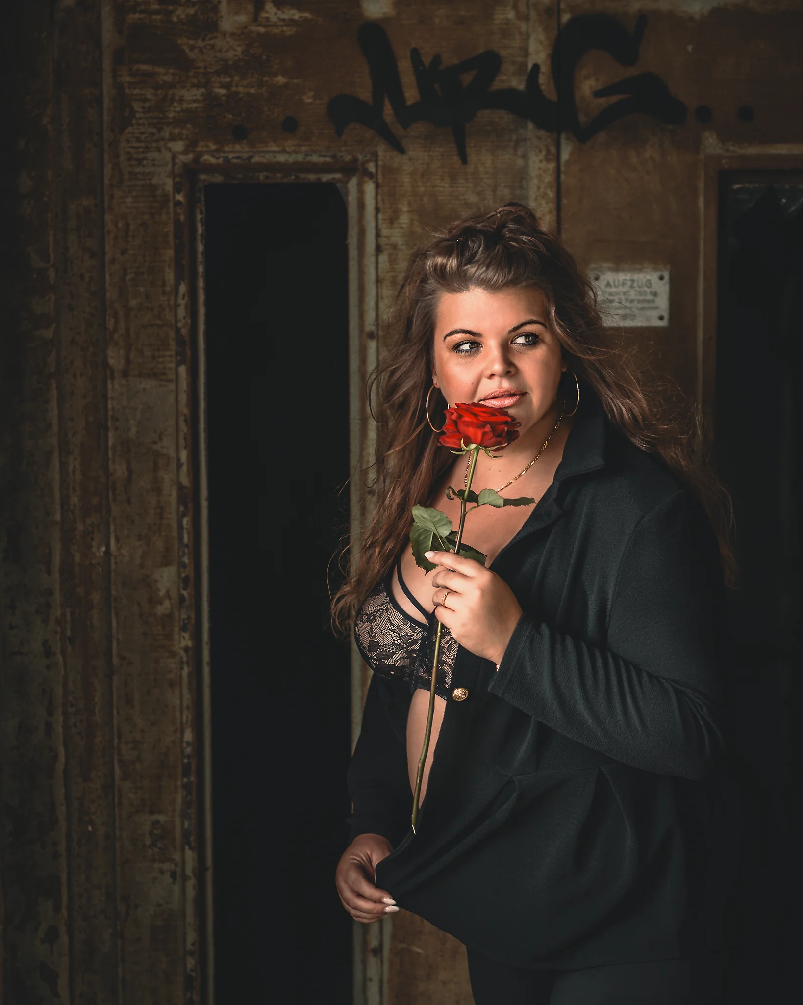 Lost Places - Porträt einer Frau vor einer rostigen, industriellen Aufzugstür mit Graffiti; sie trägt ein offenes schwarzes Outfit mit Spitzen-BH und hält eine rote Rose an den Lippen, während warmes, gerichtetes Licht ihr Gesicht betont und der raue Hintergrund eine kontrastreiche, sinnliche Atmosphäre schafft.