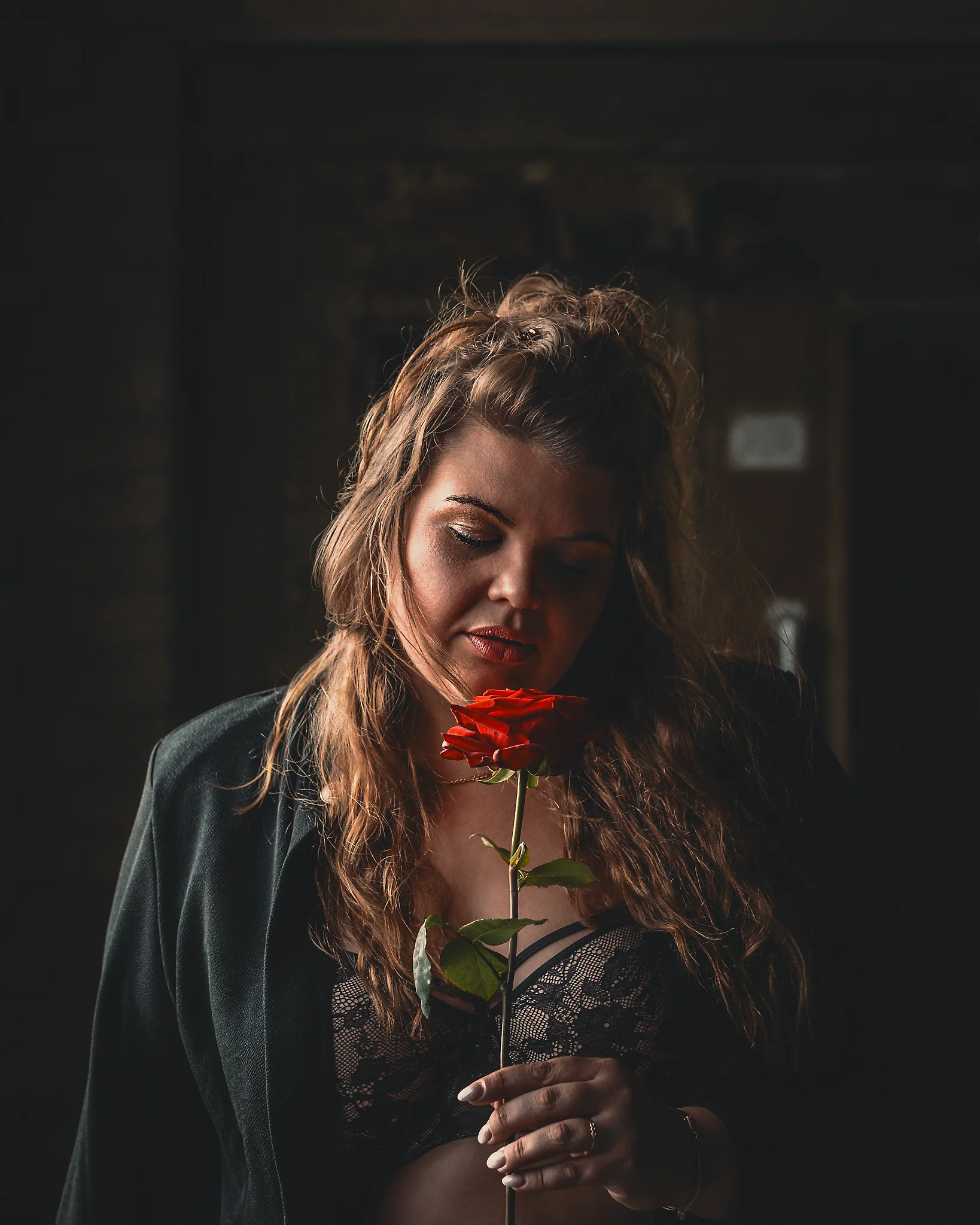 Lost Places - Stimmungsvolles Porträt einer Frau in dunkler, industrieller Umgebung; sie hält eine rote Rose vor sich und blickt nach unten, während gerichtetes, warmes Licht ihr Gesicht und die Blüte hervorhebt und der Hintergrund in weiche Schatten fällt, wodurch eine ruhige, nachdenkliche Atmosphäre entsteht.