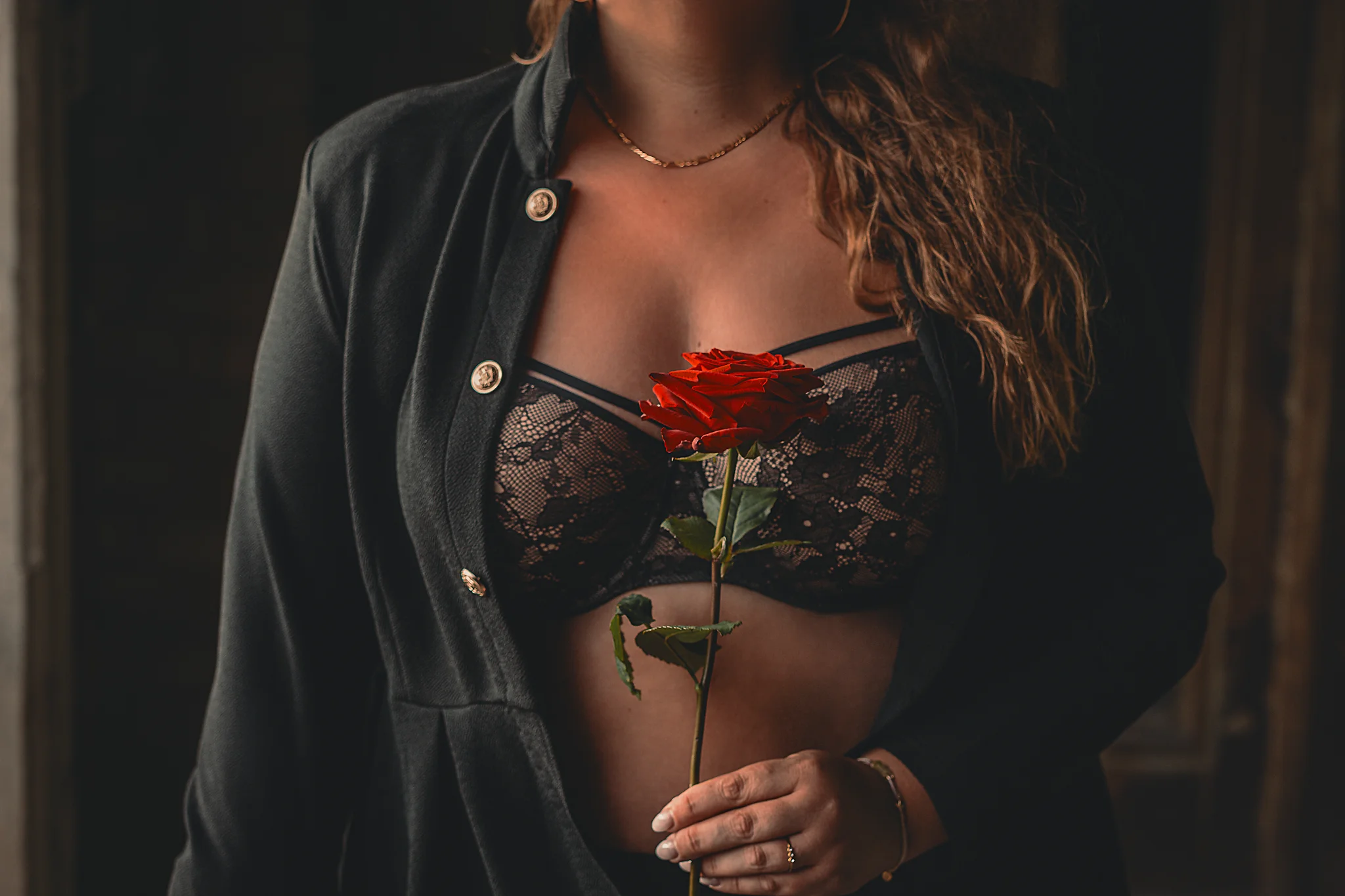 Lost Places - Detailaufnahme eines Oberkörpers in dunkler, industrieller Umgebung: Eine Frau im offenen schwarzen Blazer und Spitzen-BH hält eine rote Rose vor der Brust, während warmes, gerichtetes Licht die Blüte und die Texturen hervorhebt und der Hintergrund weich in Schatten bleibt.