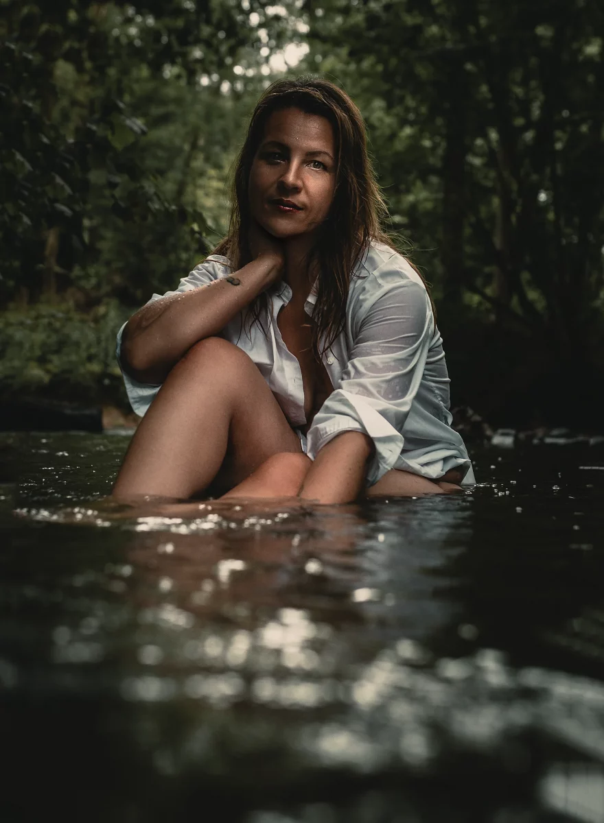 Natur - Person sitzt in einem flachen Bach im Wald, trägt ein nasses, leicht geöffnetes Hemd und legt eine Hand an den Nacken, während sie ruhig in die Kamera blickt.