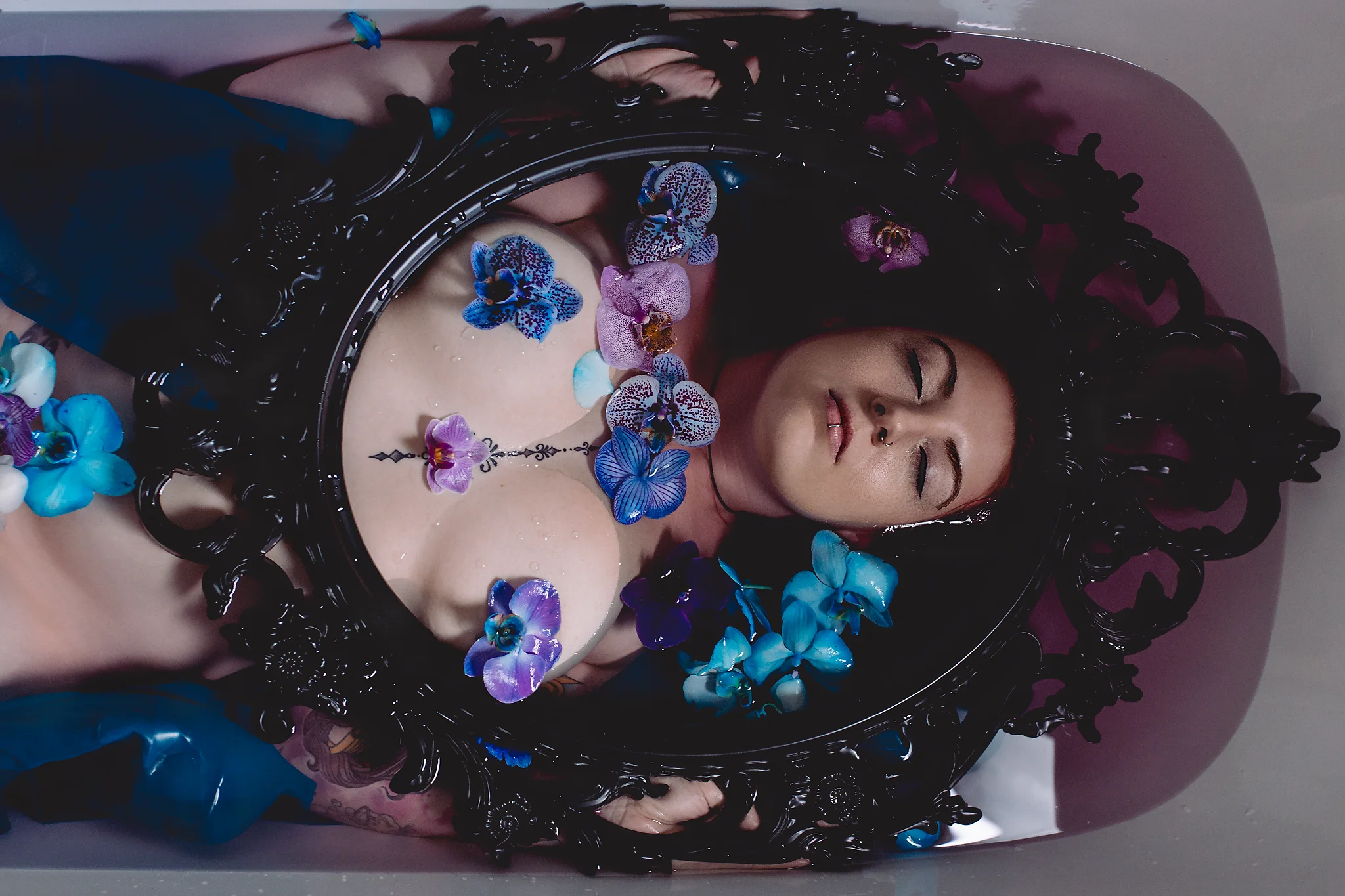 Creative - Aktporträt einer Frau, die in einer Badewanne mit leicht gefärbtem Wasser liegt; ihr Oberkörper ist von einem kunstvollen, schwarzen Spiegelrahmen eingefasst, während blaue und violette Orchideen auf ihrer Haut treiben, ihre Augen geschlossen sind und das weiche, gerichtete Licht eine ruhige, fast meditative Atmosphäre mit starkem Farbkontrast erzeugt.