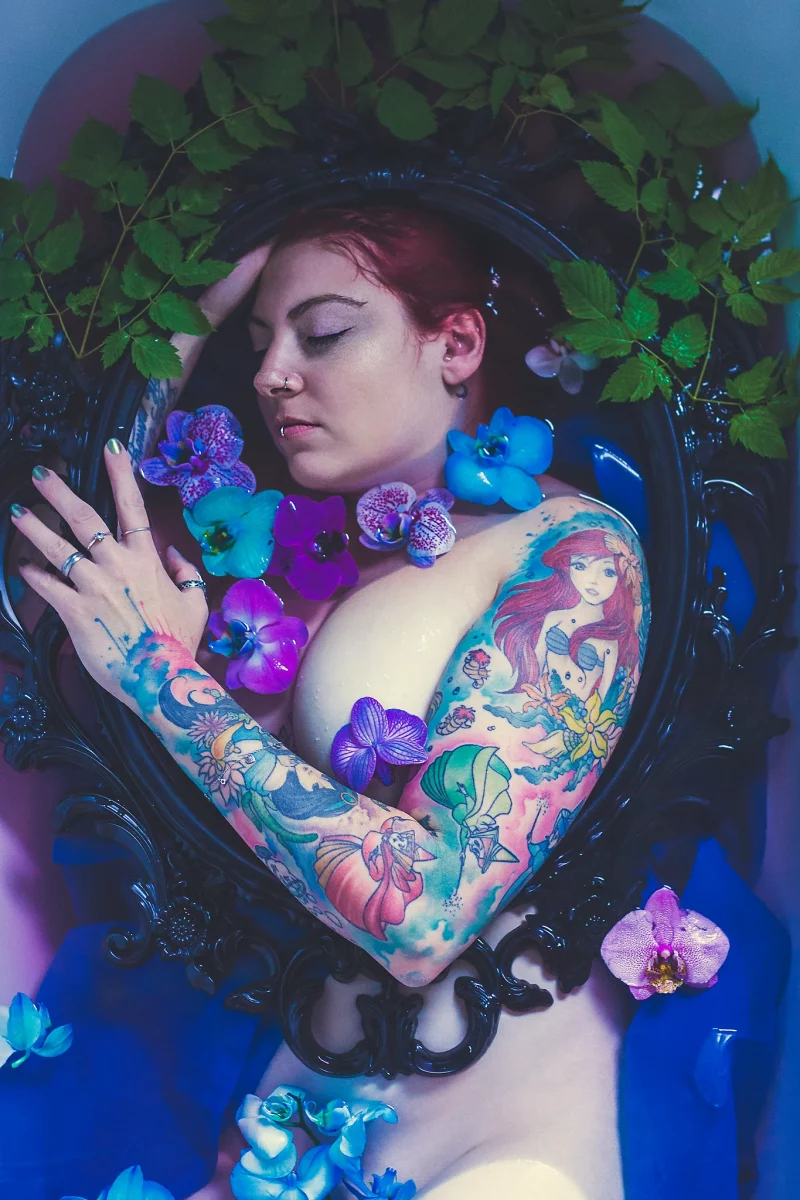 Creative - Aktporträt einer Frau mit roten Haaren, die in einer Badewanne mit farbigem Wasser liegt; ihr Oberkörper ist von einem kunstvollen schwarzen Spiegelrahmen eingefasst, während blaue und violette Orchideen sowie grüne Blätter auf dem Wasser treiben, ihre Augen geschlossen sind und das weiche, farbige Licht eine ruhige, verträumte und künstlerische Atmosphäre erzeugt.