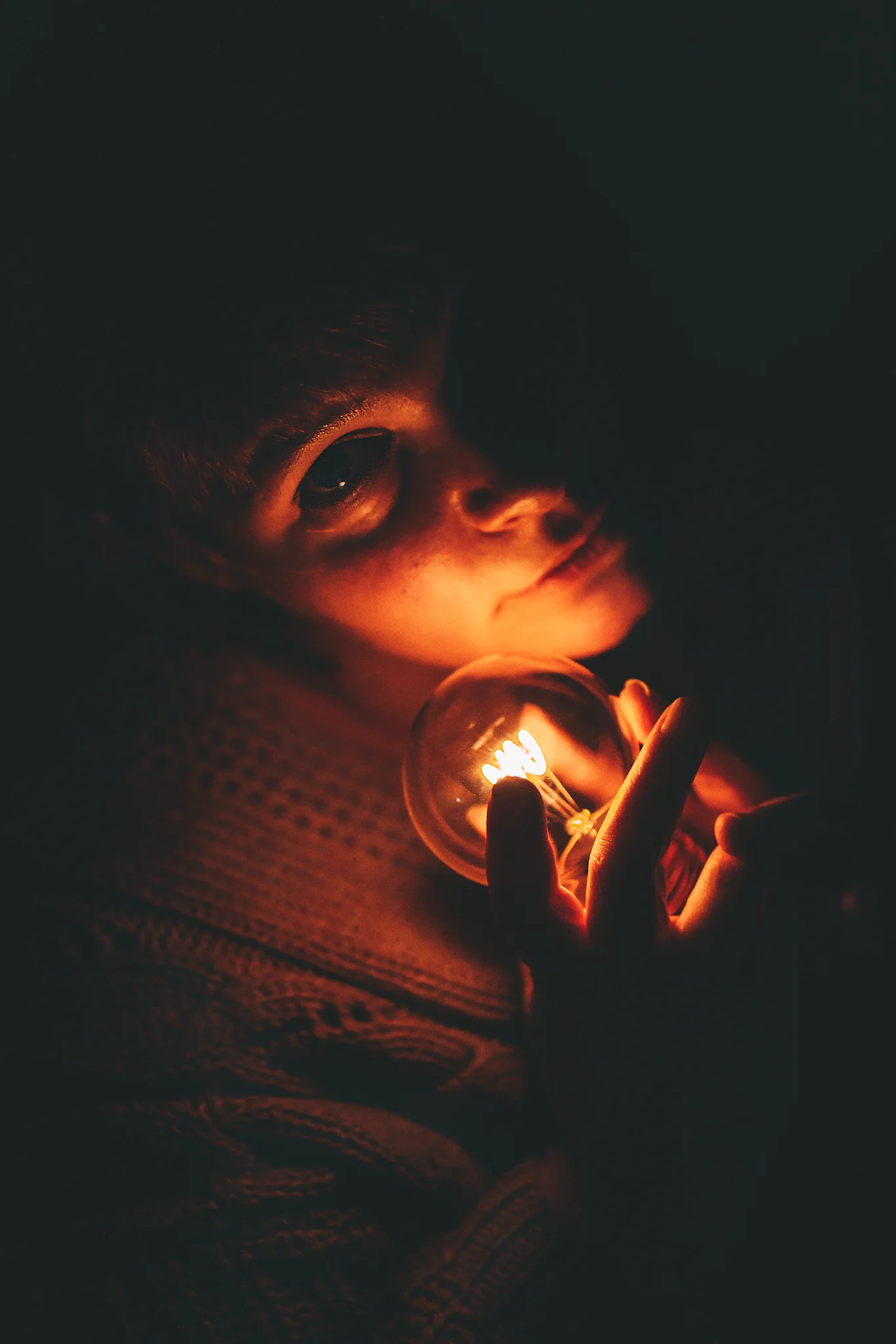 Creative - Porträt einer Frau in dunkler Umgebung, die eine leuchtende Glühbirne in den Händen hält; das warme, punktuelle Licht beleuchtet ihr Gesicht von unten, betont ihre Augen und schafft starke Schatten, wodurch eine intime, geheimnisvolle und fast cineastische Stimmung entsteht.